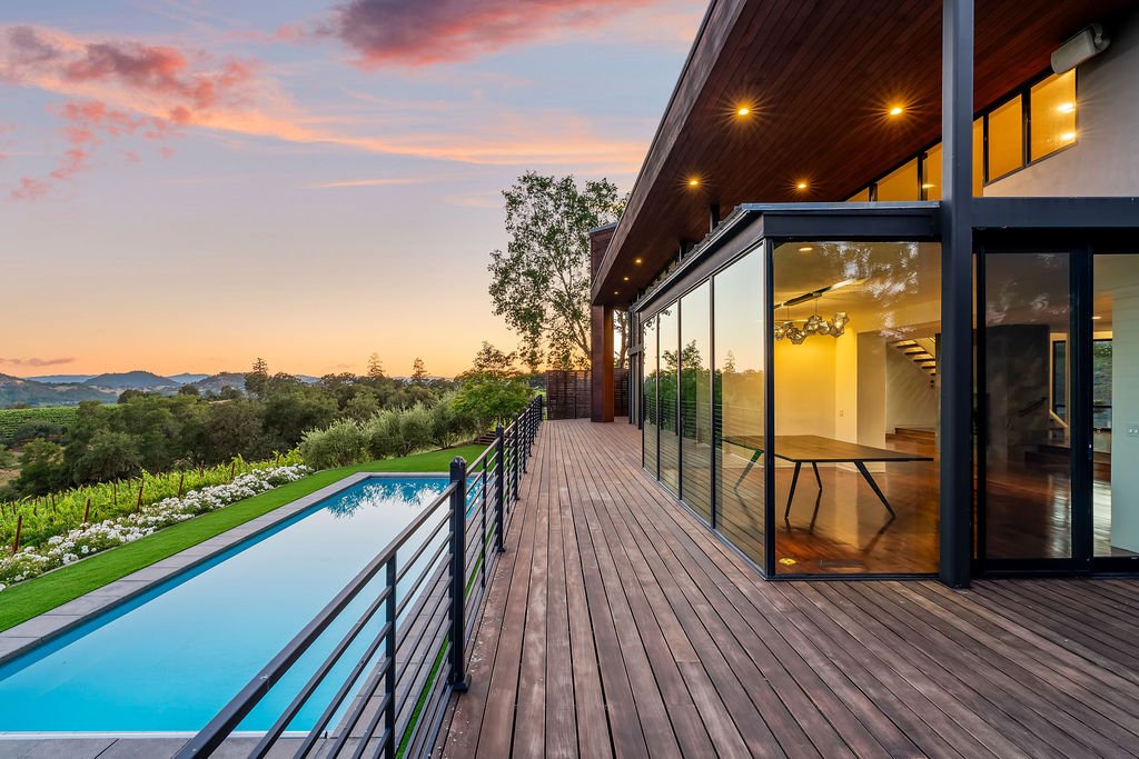 Modern house with a wooden deck, large glass windows, outdoor pool, and scenic countryside view at sunset.