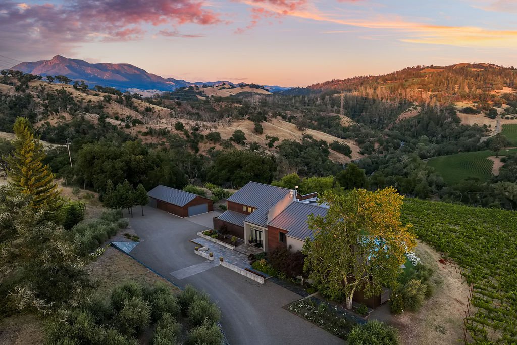 A house with a metal roof situated on a hillside with surrounding trees and fields, with mountains in the background under a colorful sky at sunset.
