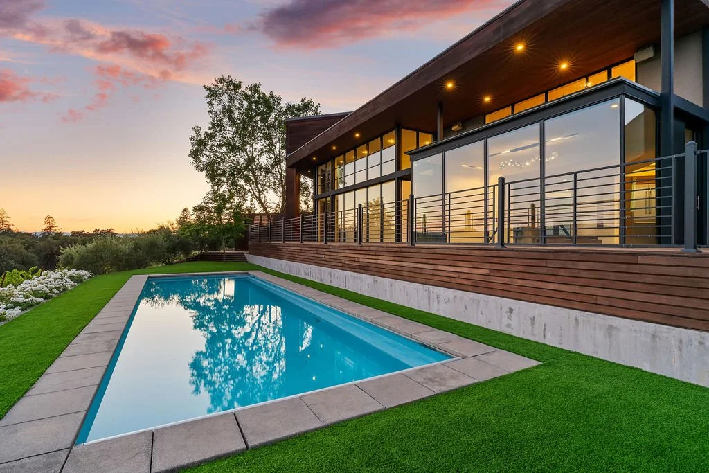 Modern house with large windows, an elevated deck, and a swimming pool surrounded by green lawn at sunset.