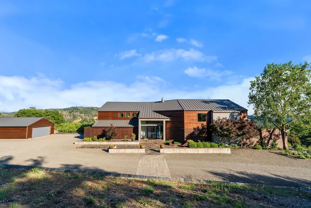 A modern, two-story house with a metal roof and wooden exterior, surrounded by a landscaped yard with trees and plants, under a blue sky.