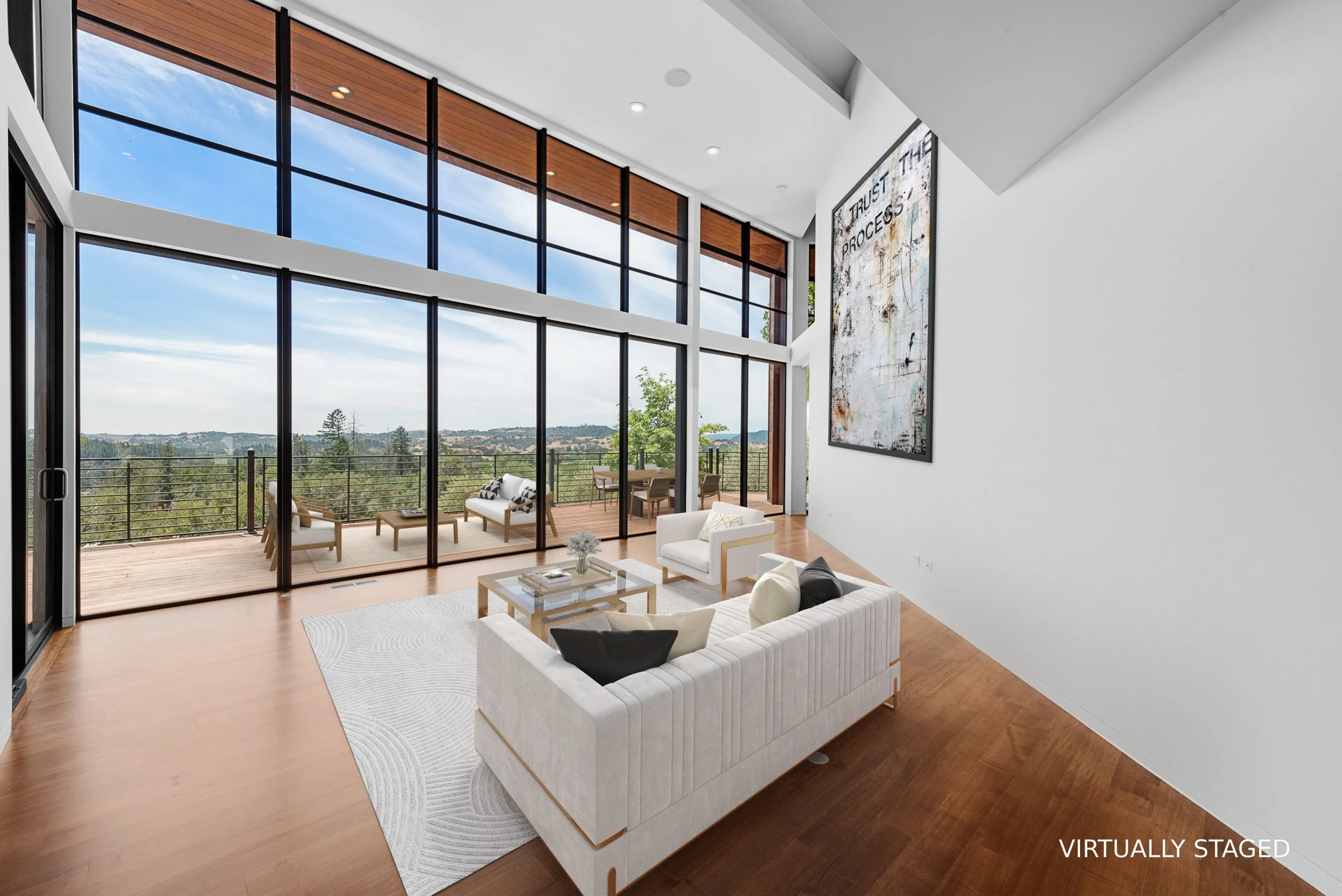 A modern living room with large floor-to-ceiling windows and sliding glass doors opening to a balcony with outdoor furniture and a scenic view of treetops and rolling hills. The room has white walls, wooden flooring, and contemporary furniture, inclu