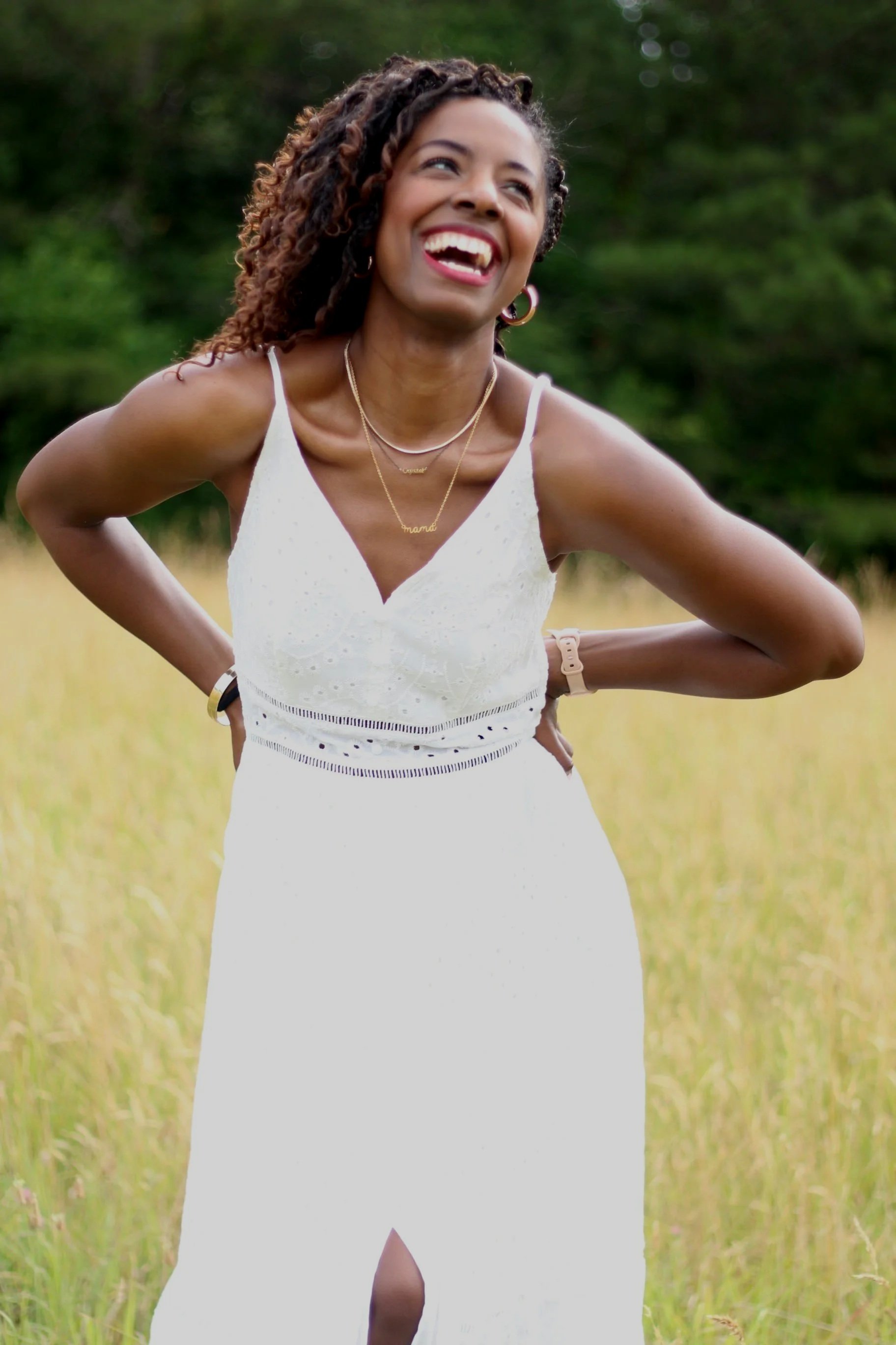 A woman in a white dress smiling outdoors in a grassy field with green trees in the background.