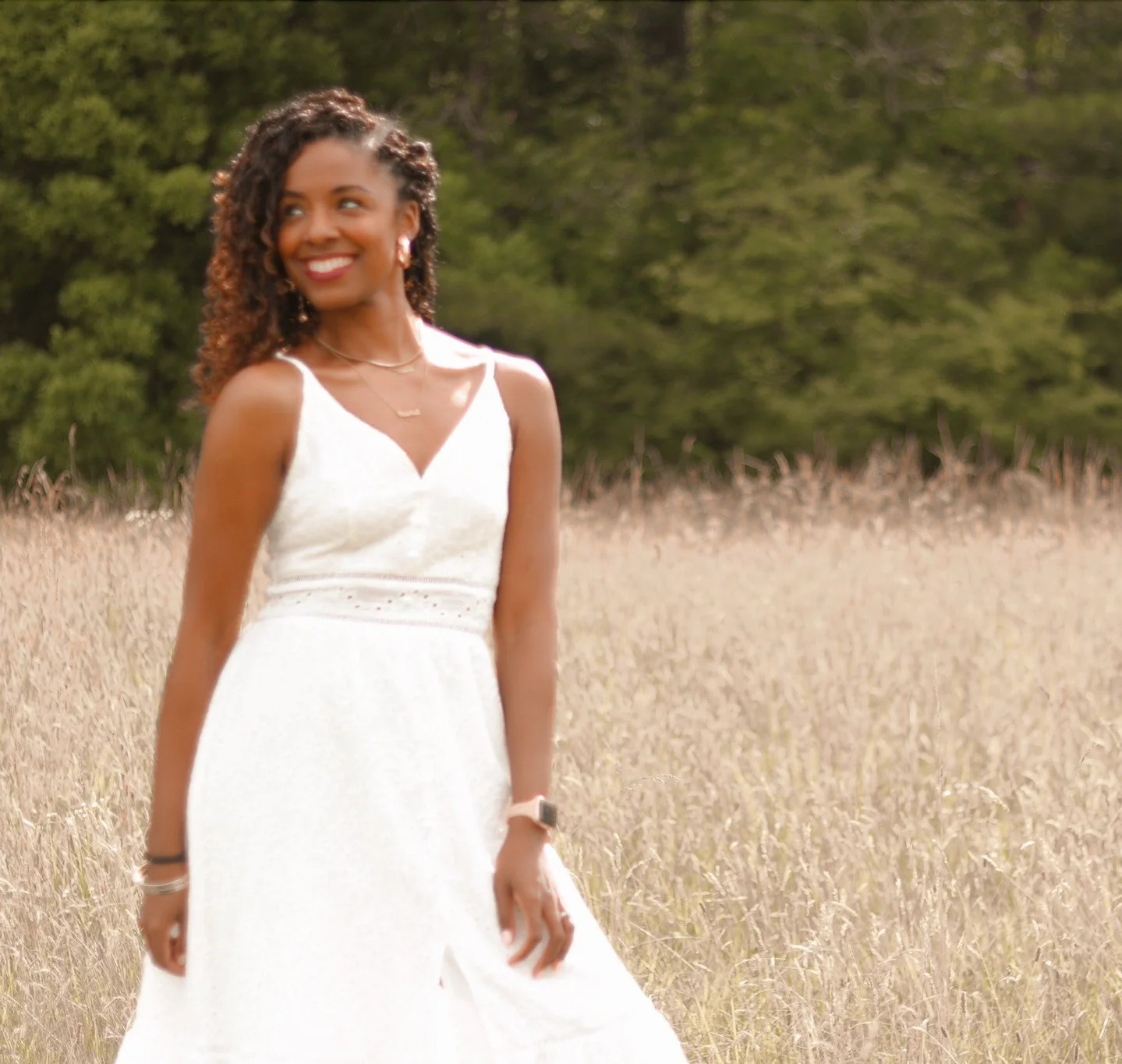 A woman in a white sleeveless dress standing in a field of dry grass with green trees in the background, smiling and looking to her left.