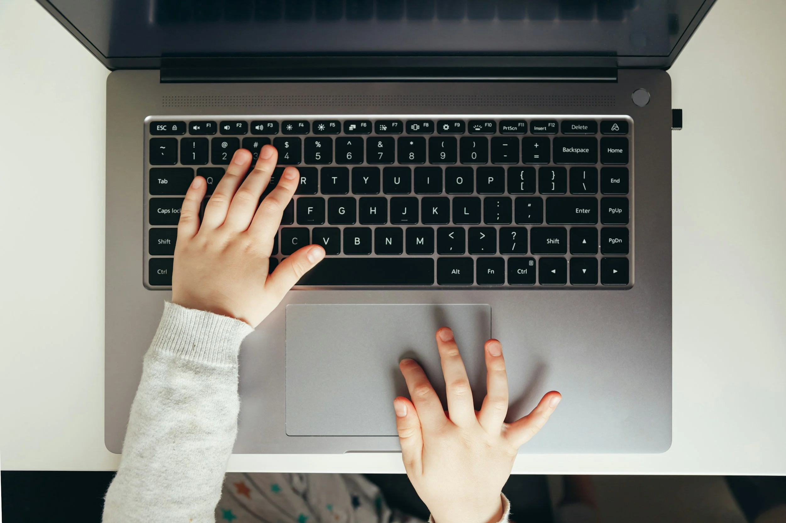 Child's hands typing on a silver laptop keyboard with a white background.