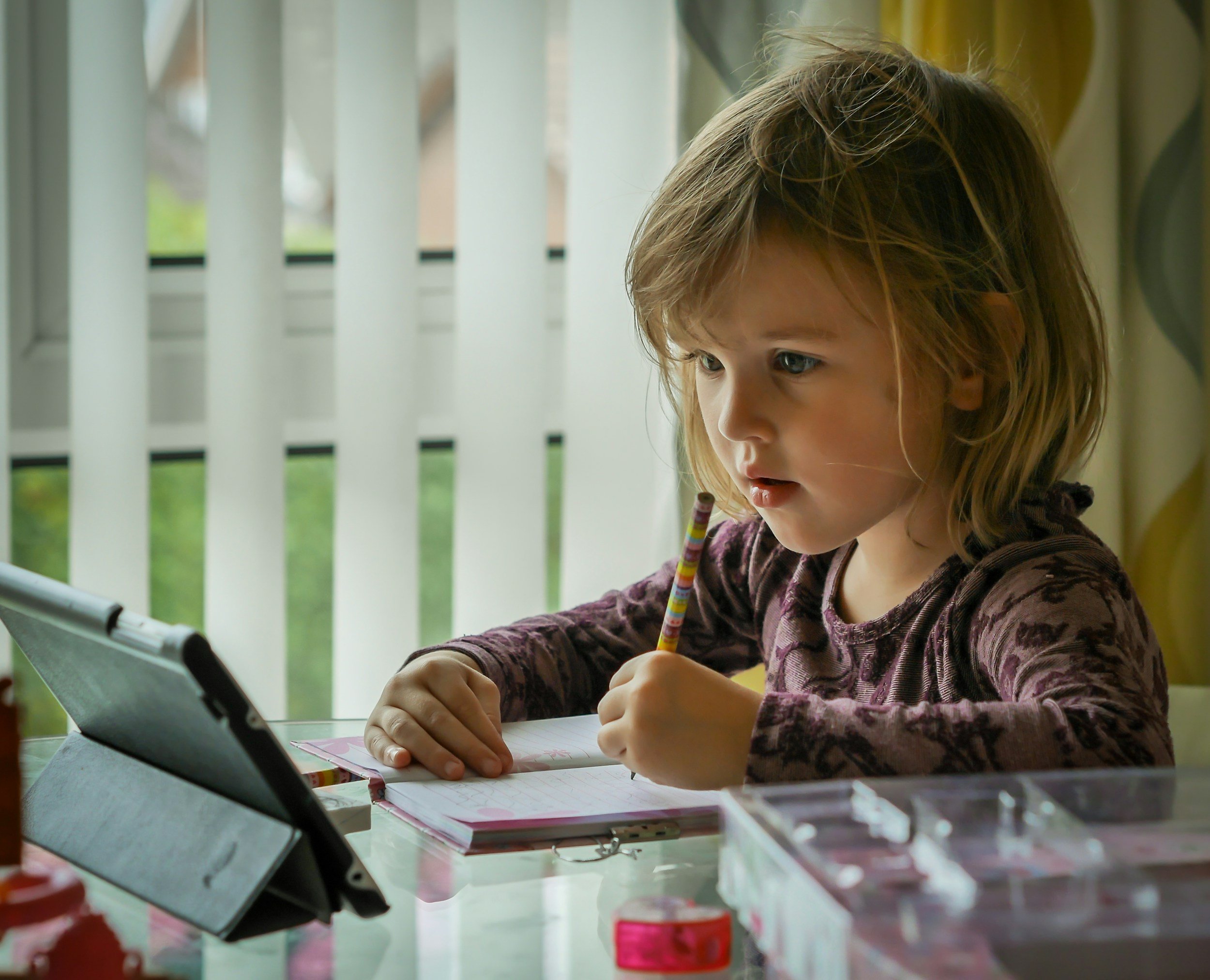 A young girl with blonde hair and blue eyes, wearing a purple patterned shirt, is sitting at a table with a tablet and a notebook, writing with a crayon. She is near a window with white blinds, and there are pink and red objects on the table.