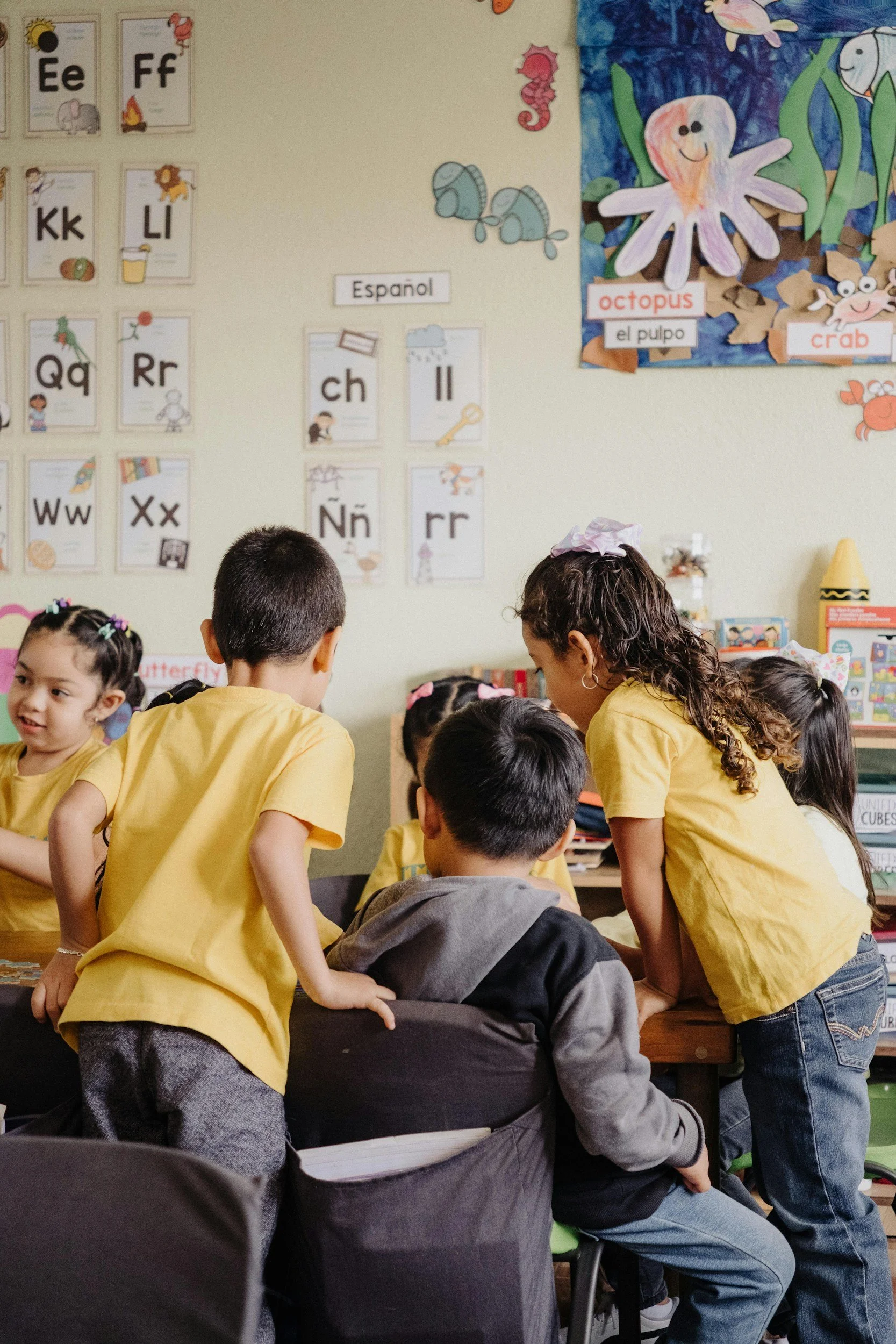 Students gathered around a classroom table, some standing and some sitting, engaging with each other. The classroom has educational posters on the wall, including the alphabet and a colorful ocean-themed display featuring an octopus, crab, and other sea creatures.