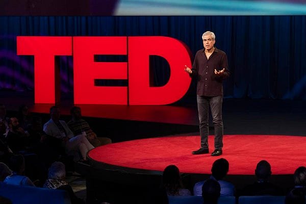 A man giving a presentation on a stage with a large red TED sign in the background and an audience seated in front.