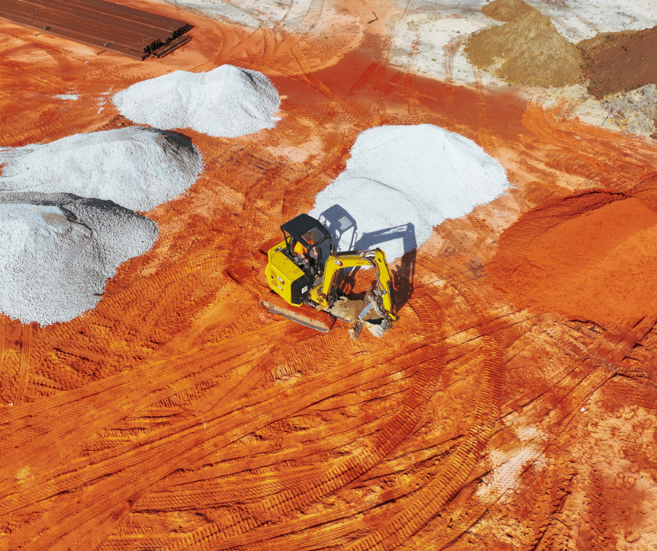 An aerial view of a construction site with a yellow excavator working on red dirt, surrounded by piles of white and gray material.