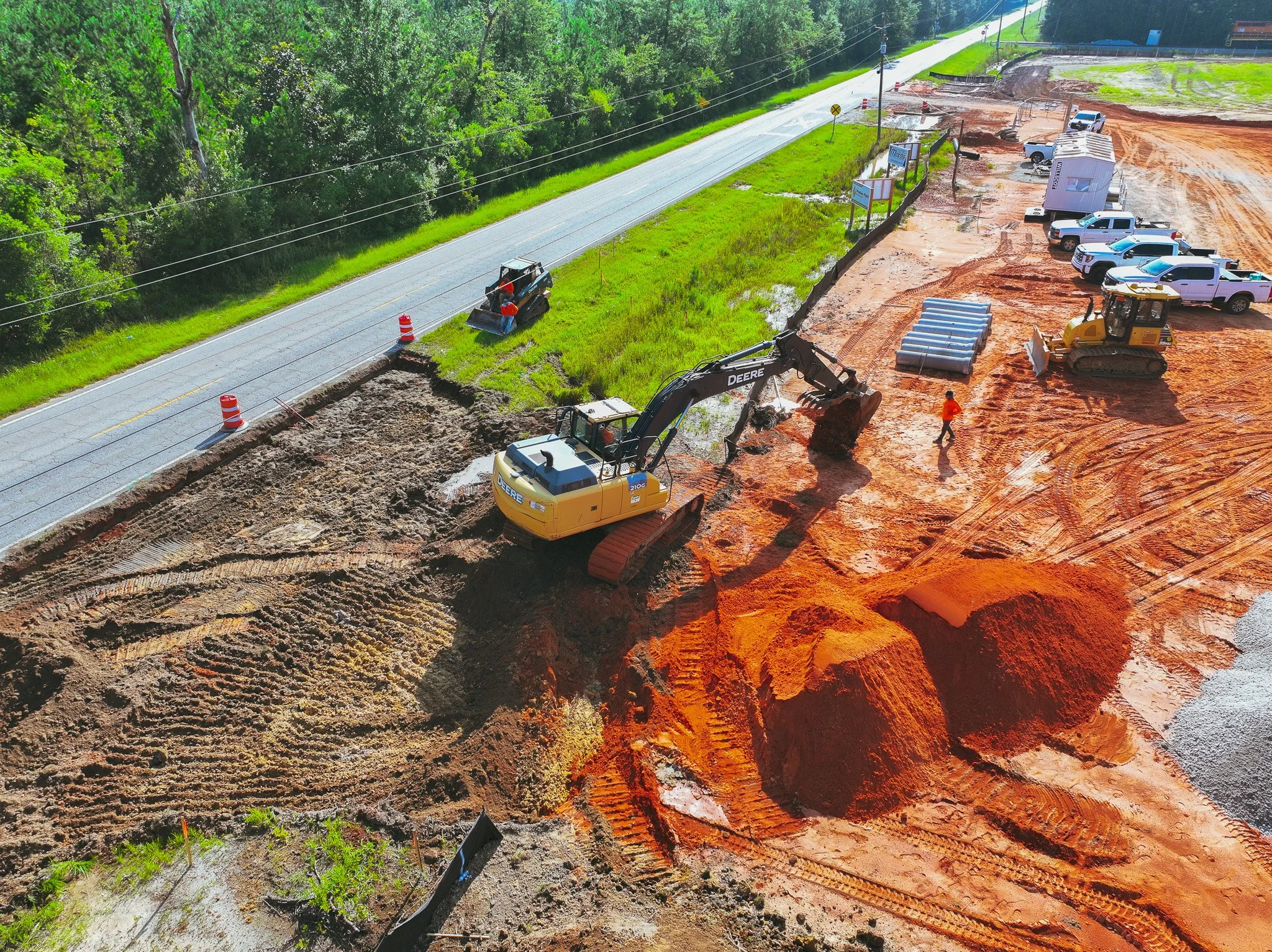 Construction site with heavy machinery and vehicles working on a road expansion project, dirt and mud on the ground, surrounded by green trees.