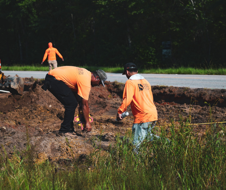 Two men working at a construction site near a road, wearing orange safety shirts, with one holding a water bottle and the other bending down. In the background, a person in an orange shirt is walking away, and there is a large excavator and green trees.
