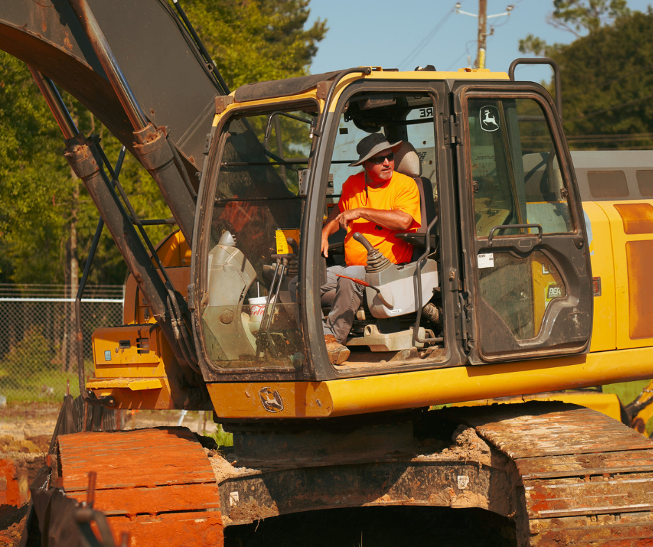 A construction worker in an orange shirt and gray pants sit in the operator's seat of a yellow excavator, looking to the side, with trees and a fence in the background.