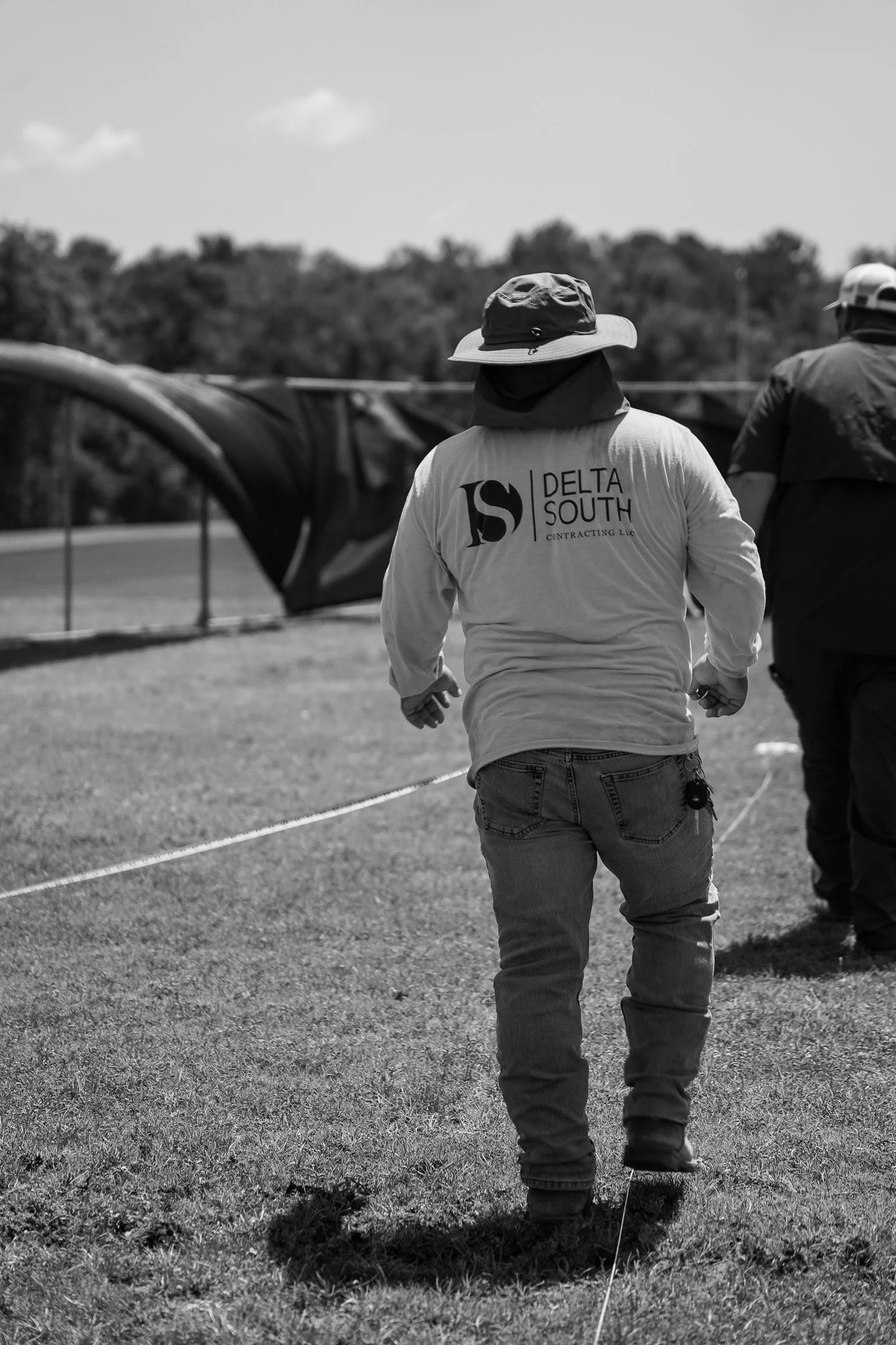 Two workers, one with a bucket hat and the other with a cap, walk along a grassy field. One wears a shirt with a business logo, and the other wears a jacket and cap. In the background, there is blurred fencing and trees.
