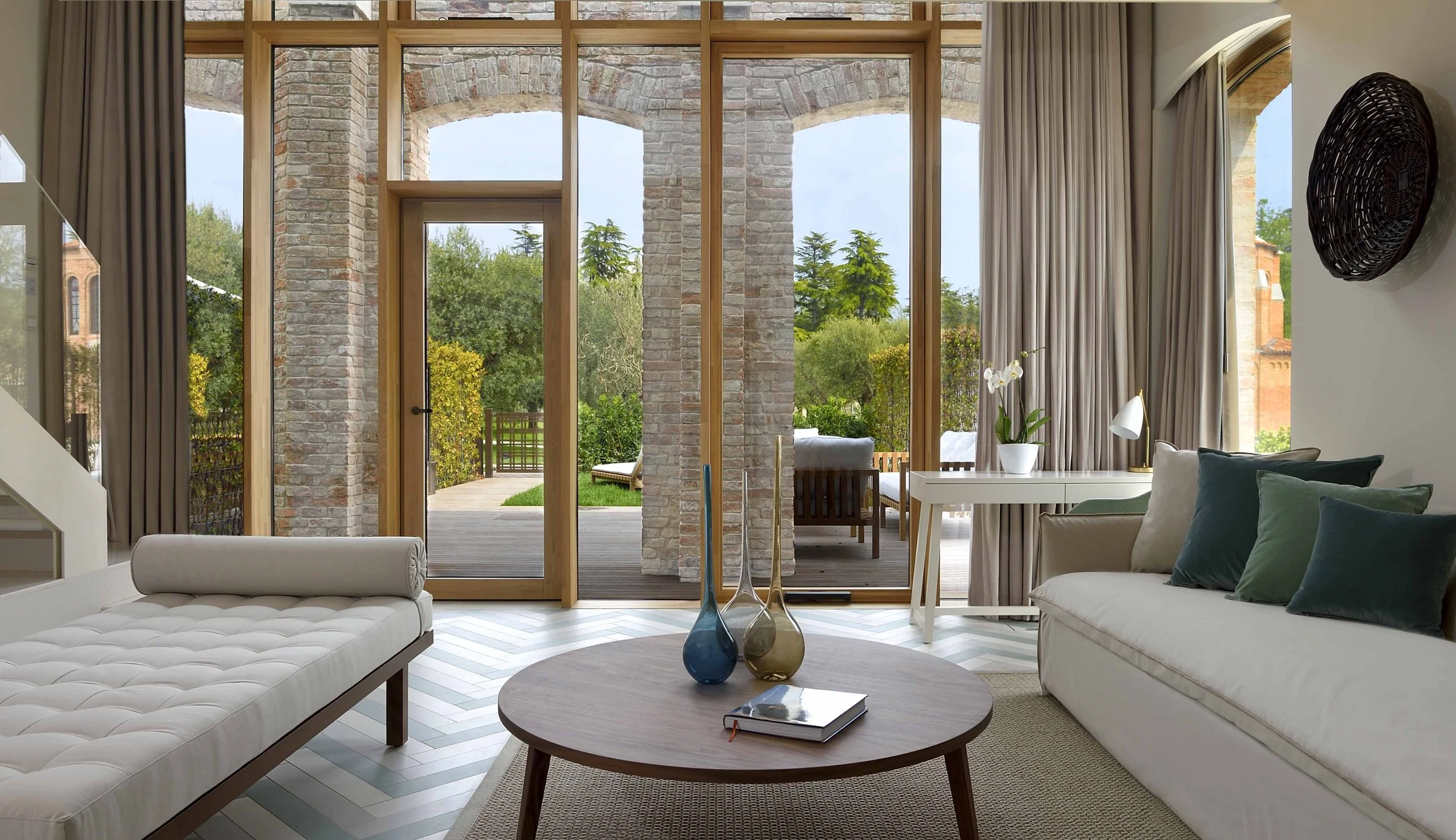Living room with large glass doors opening to a patio, brick arches, trees outside, beige curtains, white sofa with pillows, a white table with a orchid plant, vases, and a lamp, and a modern design interior.