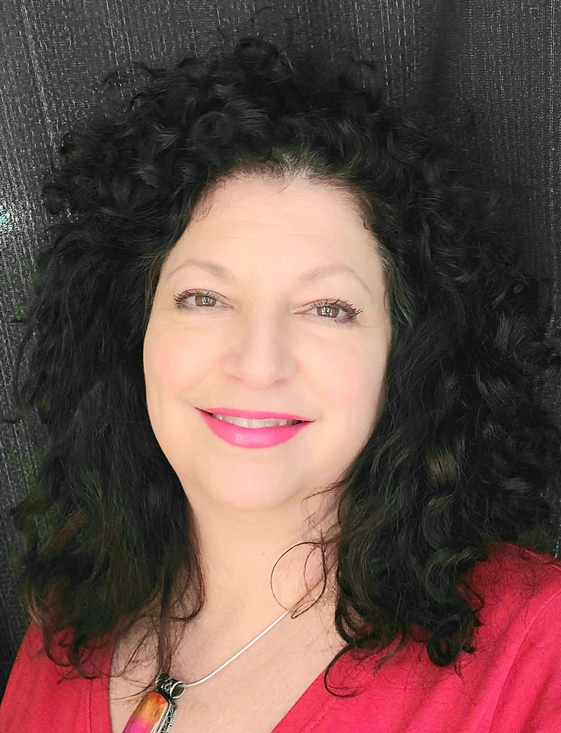 A woman with curly black hair wearing a red top with a zipper detail and a necklace, smiling and lying on a black textured surface.