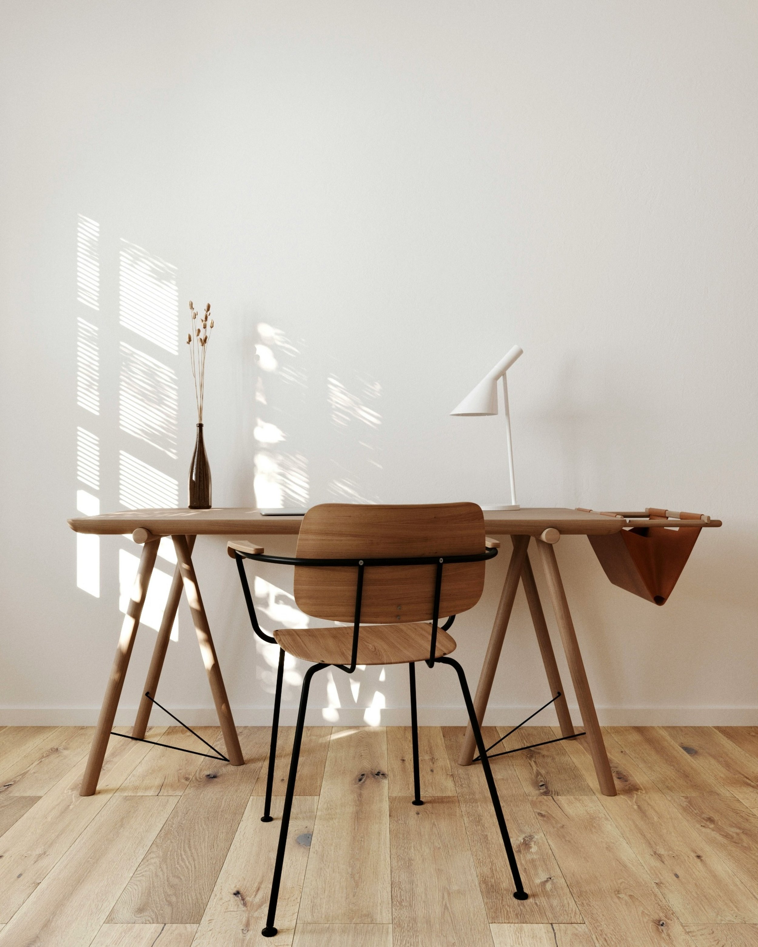 A wooden dining table with wooden chairs, a white desk lamp, a glass vase with dried flowers, and a wooden bowl on the floor, against a white wall with sunlight shadows.