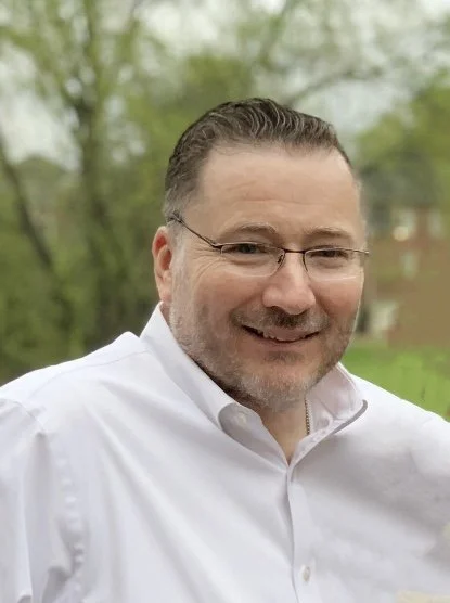 A smiling man with glasses and a beard, wearing a white shirt, outdoors with trees in the background.