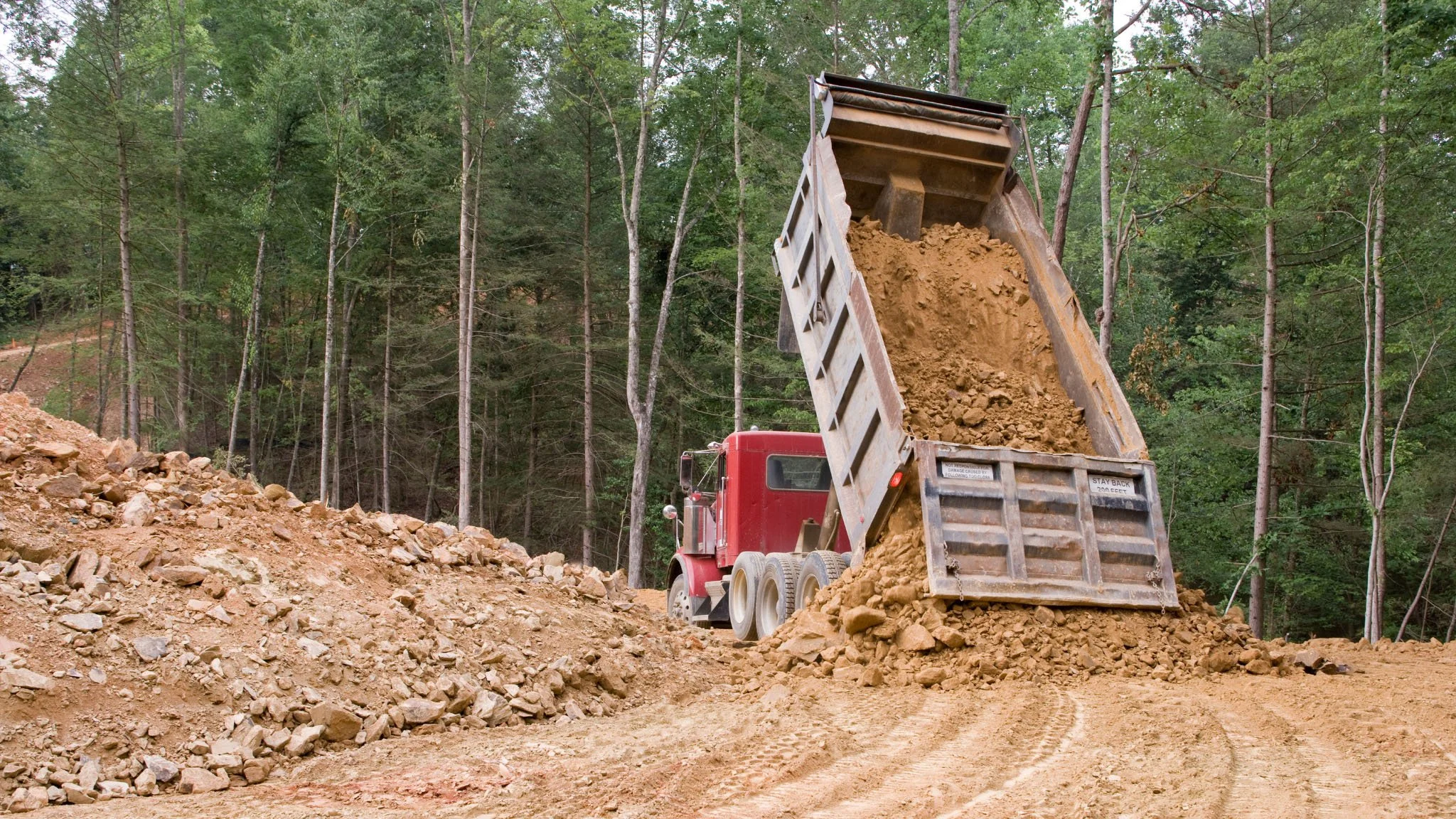 A large red dump truck unloading dirt onto a construction site surrounded by a forest of green trees.