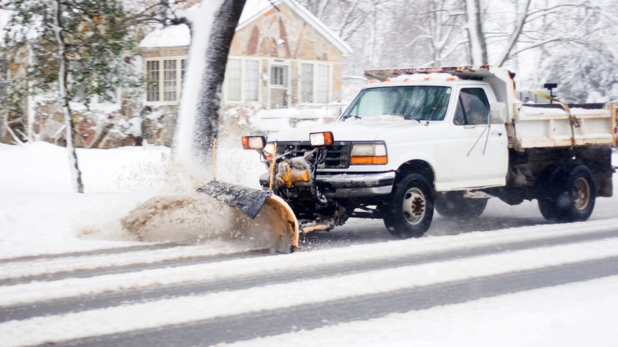 A snow plow truck clearing snow from a residential street with a house in the background.