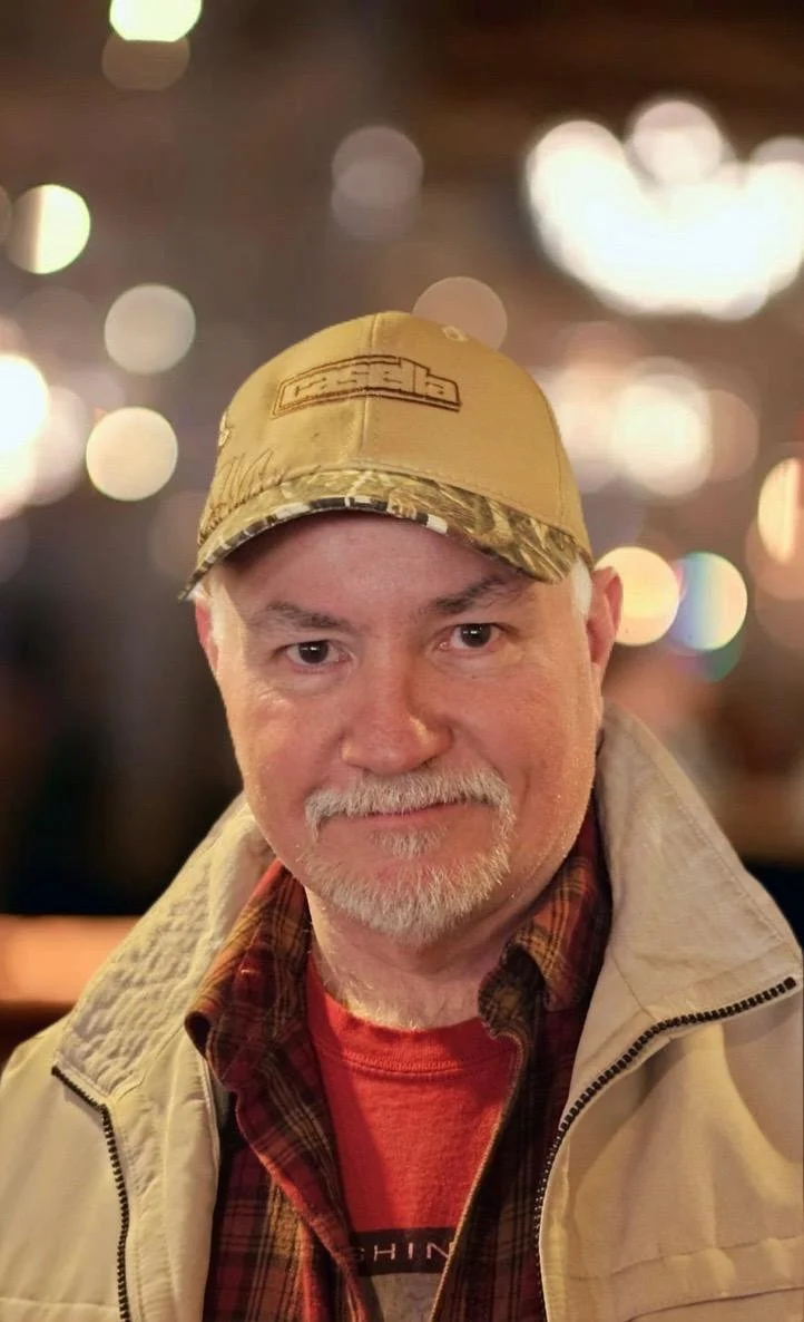 A middle-aged man with a gray beard and mustache, wearing a tan baseball cap, a plaid shirt, a red T-shirt, and a light beige jacket, smiling in an indoor setting with blurred lights in the background.