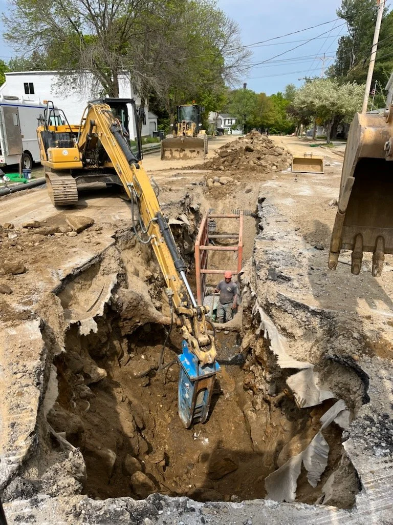 Construction workers excavating a deep trench on a residential street with excavators and dirt piles, surrounded by trees and houses.