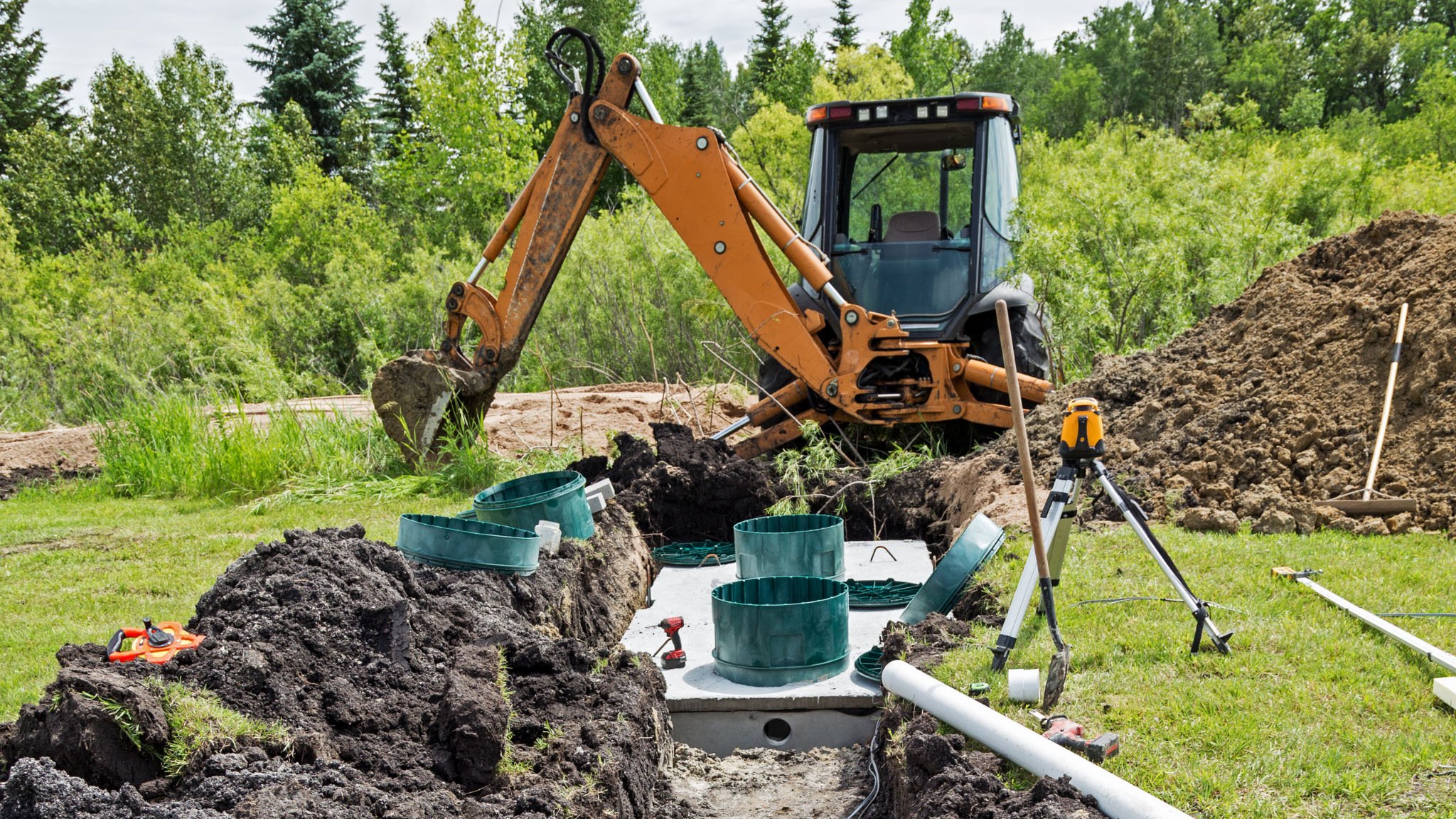 Construction site with a small orange excavator digging in the ground surrounded by green trees, with construction tools and equipment nearby.