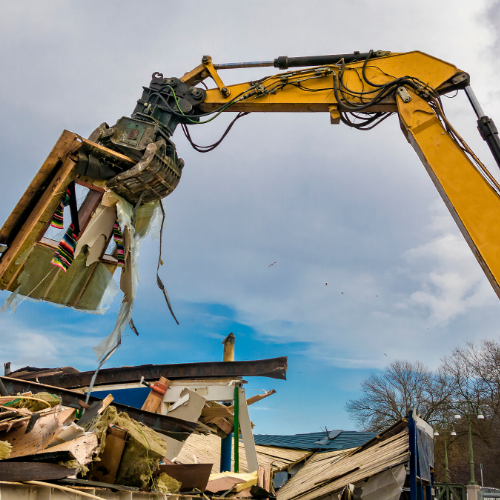 A yellow excavator demolishing a building with debris falling during the daytime.