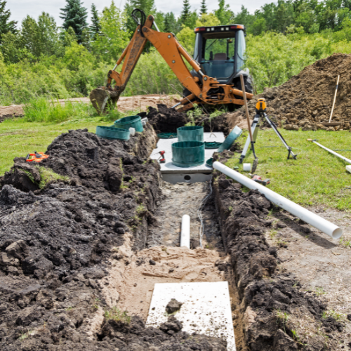 A construction site with a backhoe excavator digging trenches for underground pipe installation, with pipes and equipment nearby, surrounded by green trees against a blue sky.