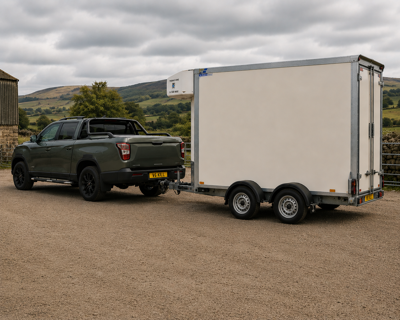 A black pickup truck towing a large white enclosed trailer on a gravel lot with rolling hills and cloudy sky in the background.