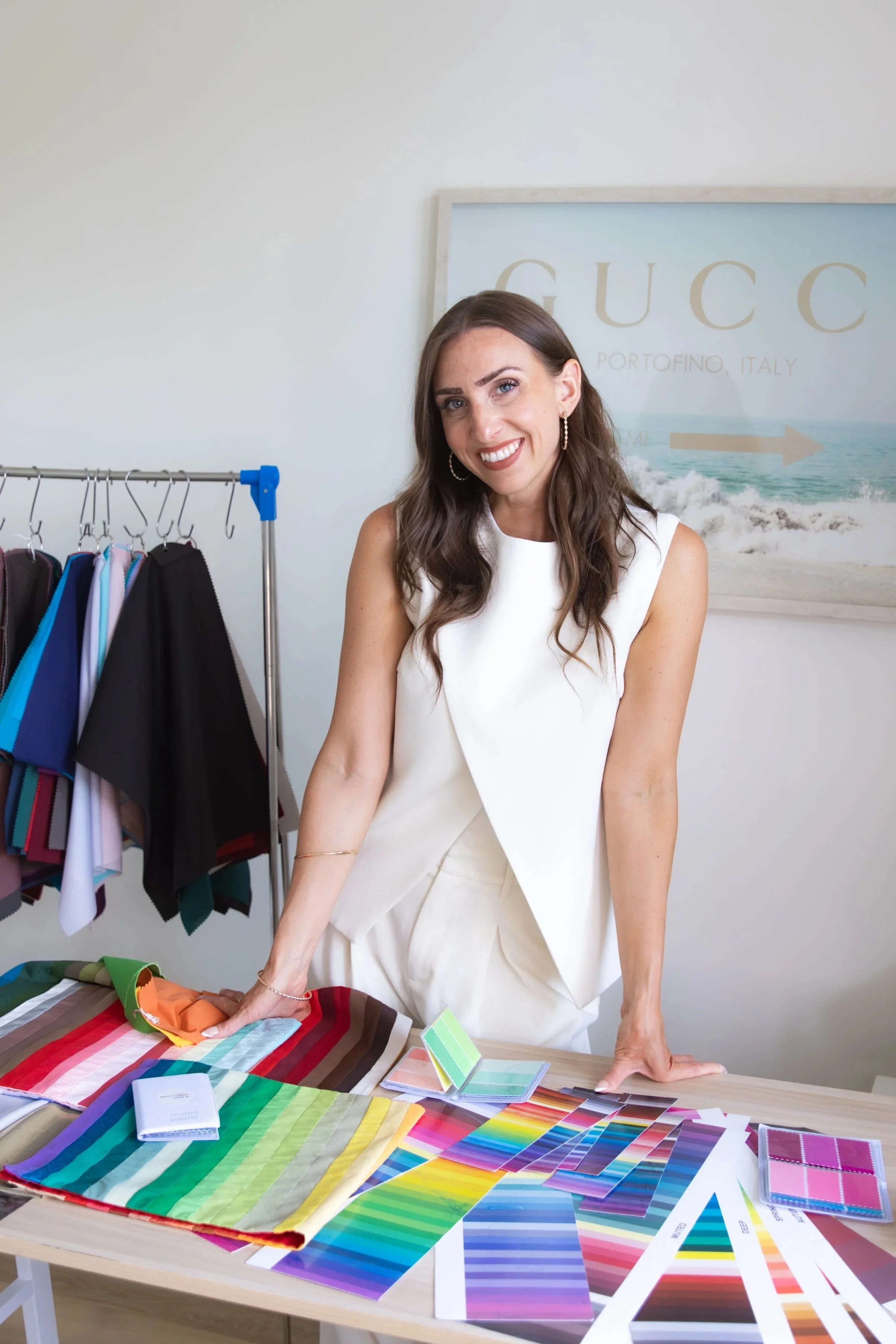 A woman smiling at a table covered with color swatches and fabric samples, with a clothing rack and a framed "GUCCI" poster in the background.
