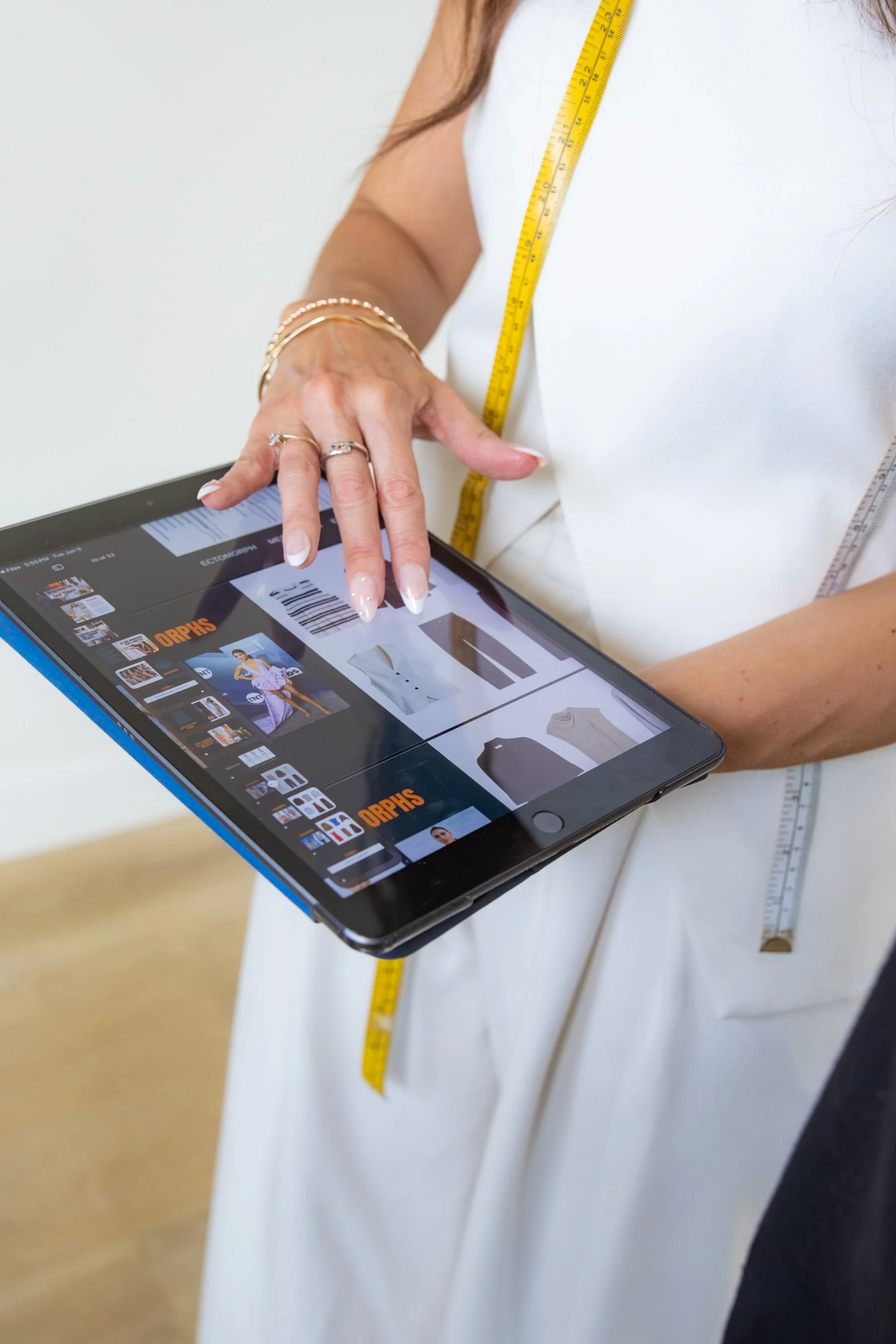 Close-up of a person wearing a white dress with a yellow measuring tape around their neck, holding a tablet displaying an online fashion store, with a focus on the person's hand. The person is wearing multiple rings and bracelets.