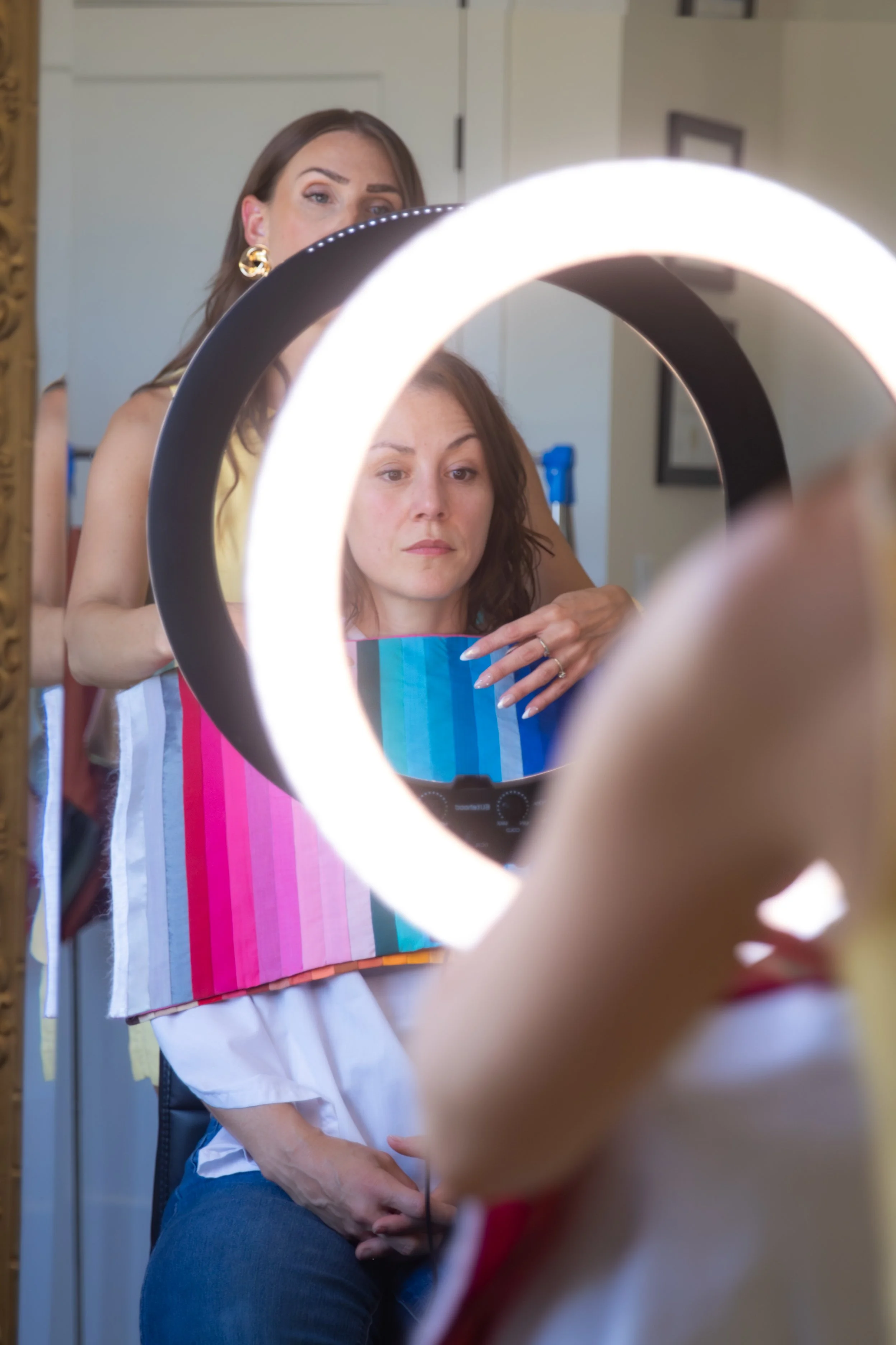 A woman wearing a beige and pink striped apron, seated in front of a mirror with ring light, looking at her reflection while getting makeup checked by another woman with gold earrings, in a room with white walls and framed pictures.