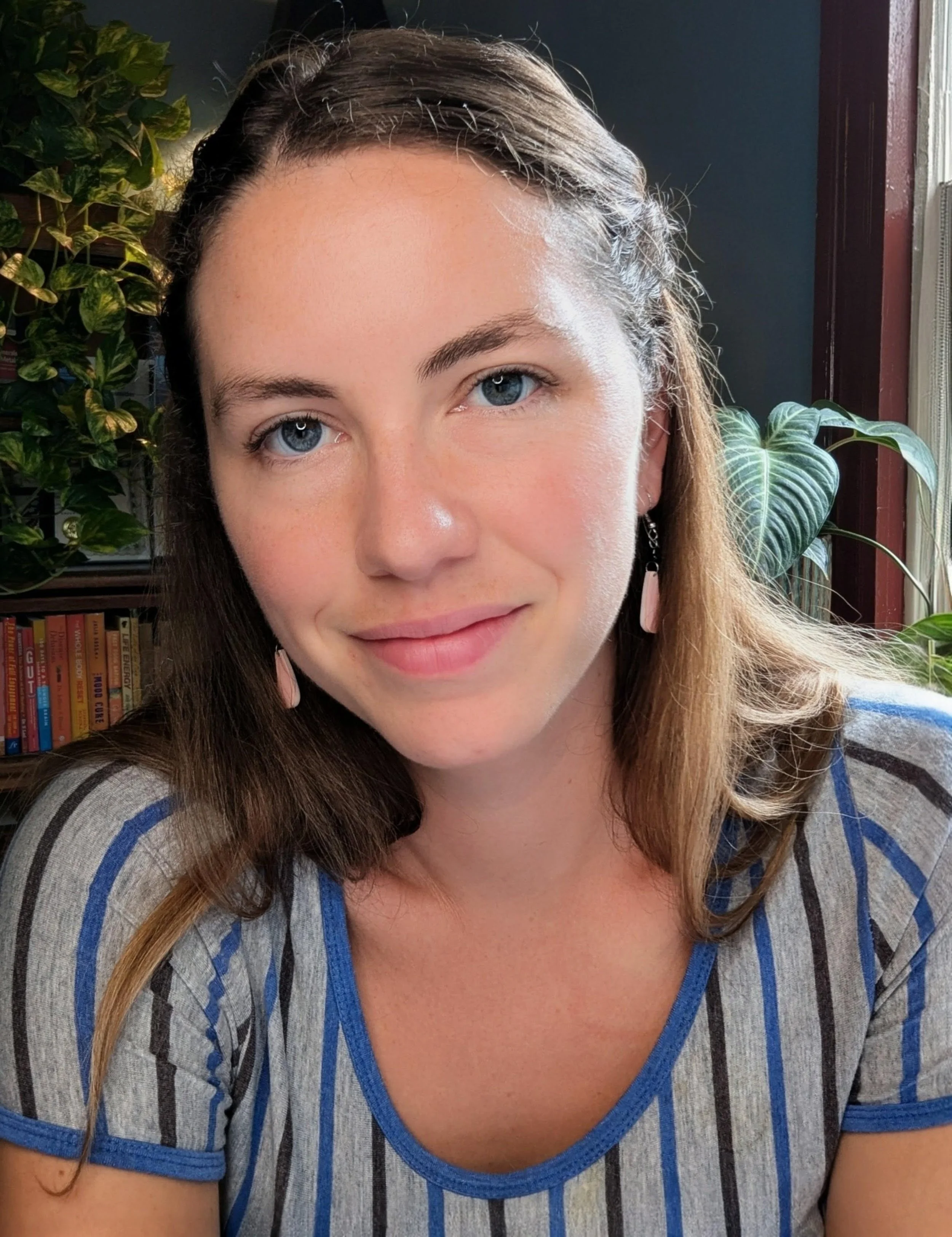 A woman smiling, with brown hair and blue eyes, wearing a gray shirt with blue trim, sitting in a room with green plants and a bookshelf in the background.
