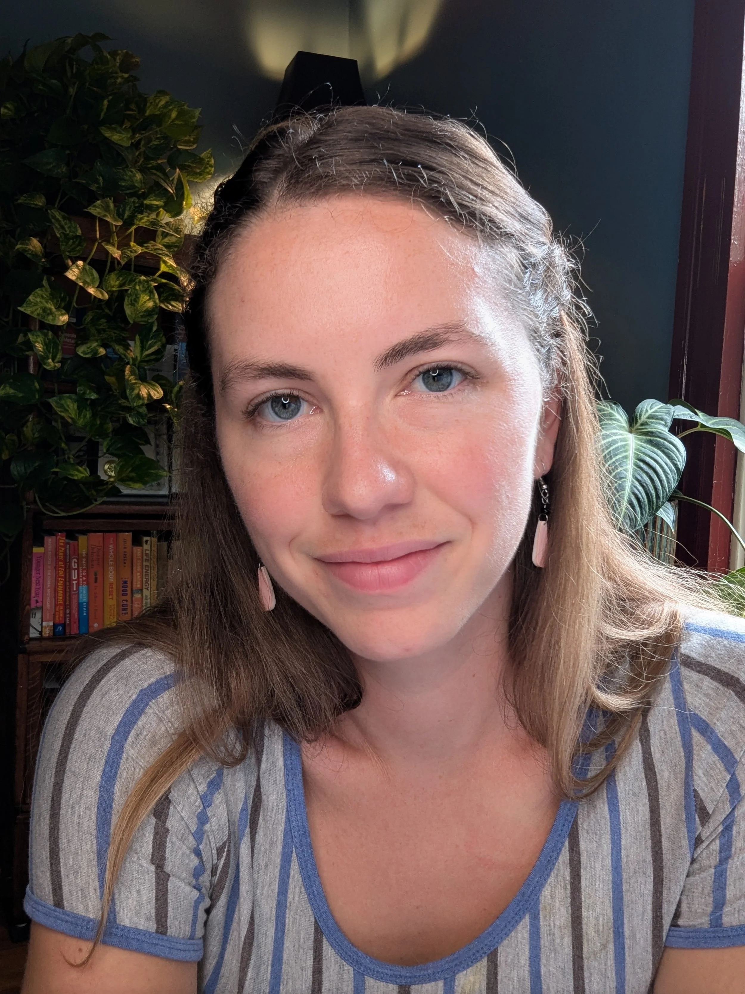 A woman with light skin and blue eyes, smiling slightly, wearing a striped shirt, sitting indoors beside a green leafy plant, with books visible in the background.