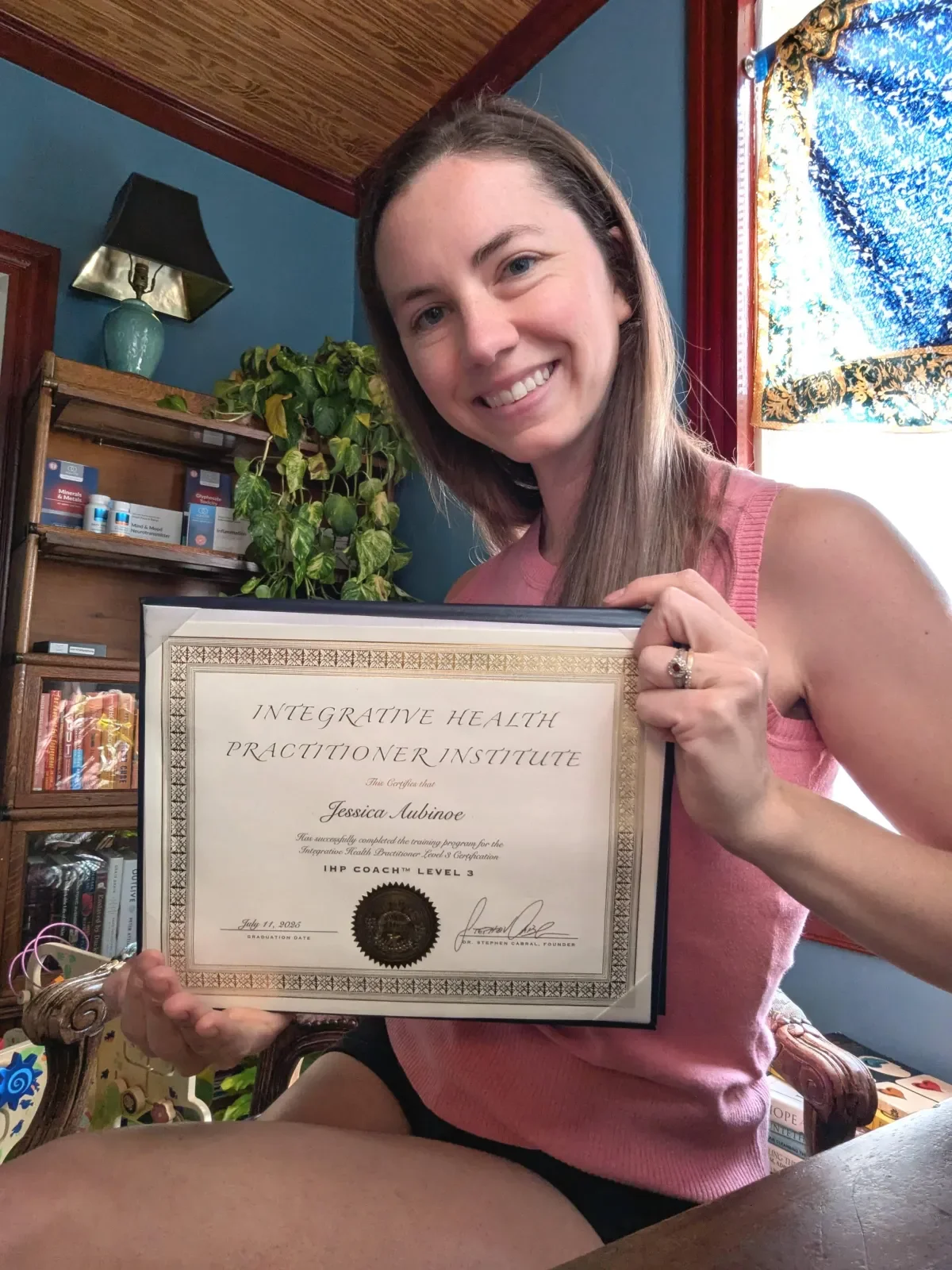 Smiling woman holding a framed certificate from the Integrative Health Practitioner Institute, sitting in a room with bookshelves, plants, and a window with colorful curtains.