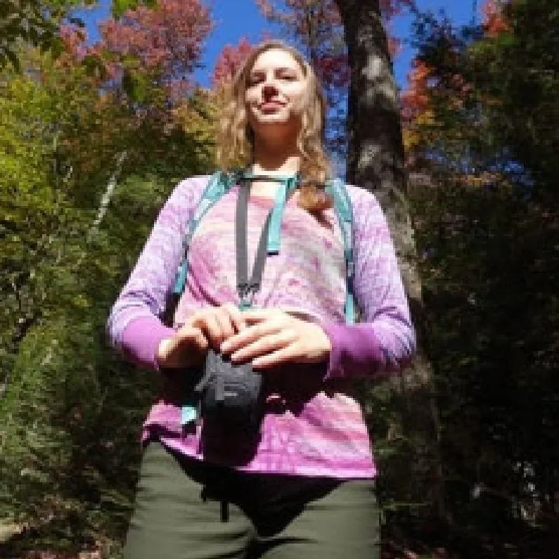 A woman standing outdoors among trees with autumn foliage, wearing a colorful long-sleeve shirt, a backpack, and holding a camera.