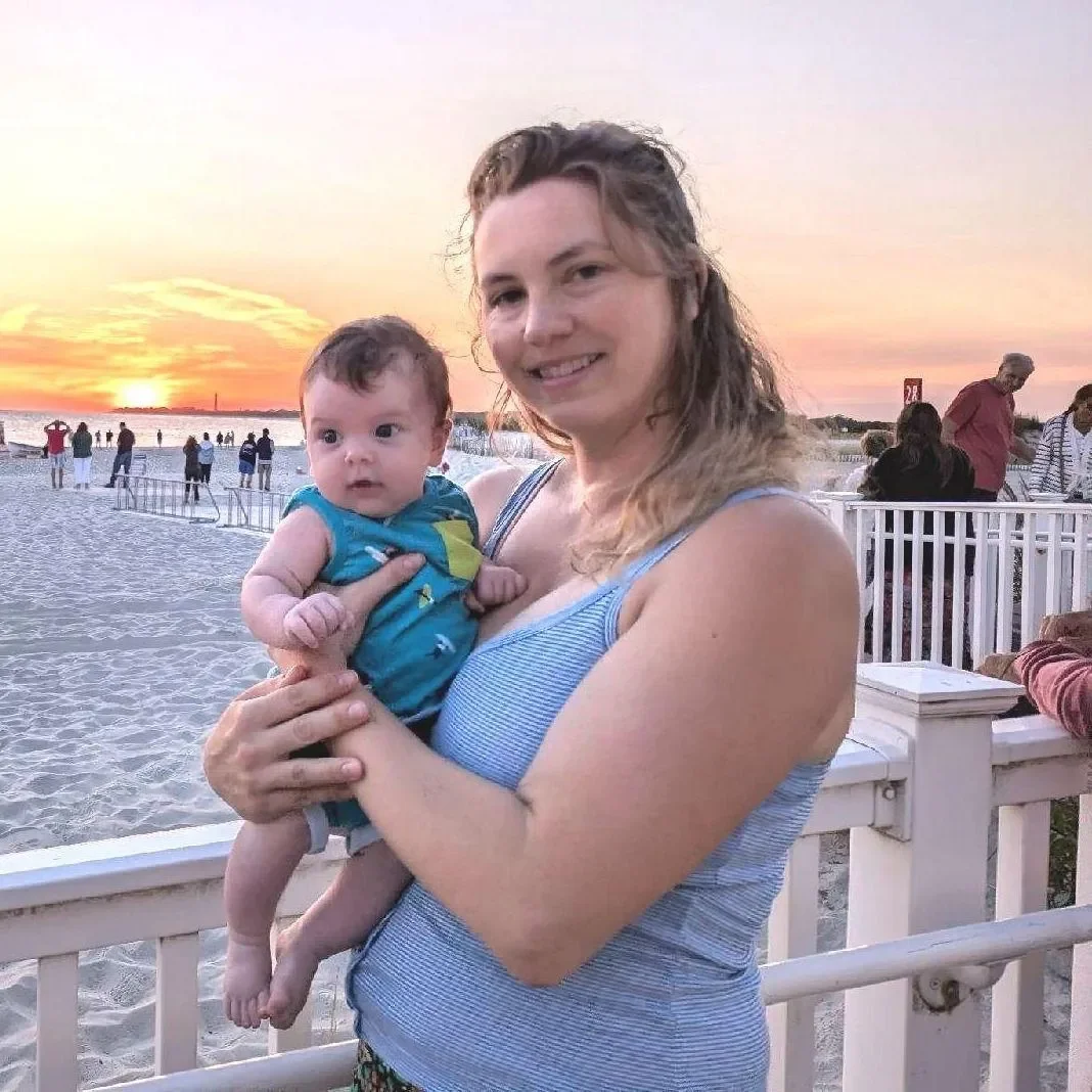A woman holding a baby on a beach at sunset with people in the background.