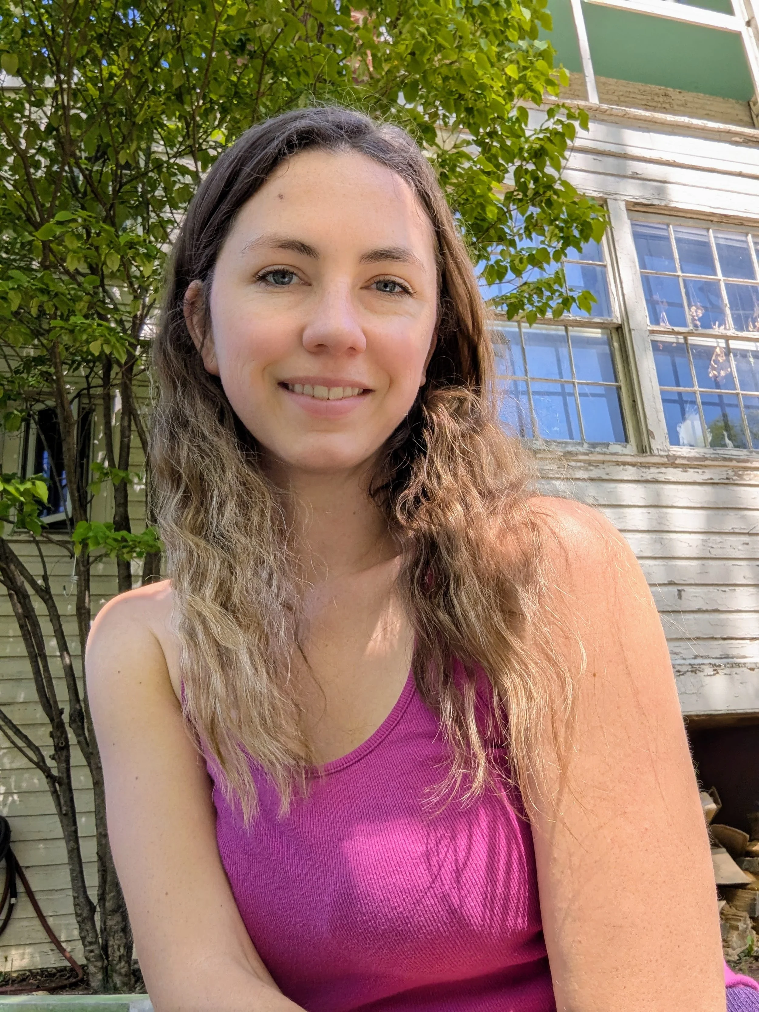 A young woman with long, wavy hair and light skin and blue eyes smiling outdoors. She is wearing a pink sleeveless top. In the background, there are green leafy trees and a peeling white wooden building with windows.