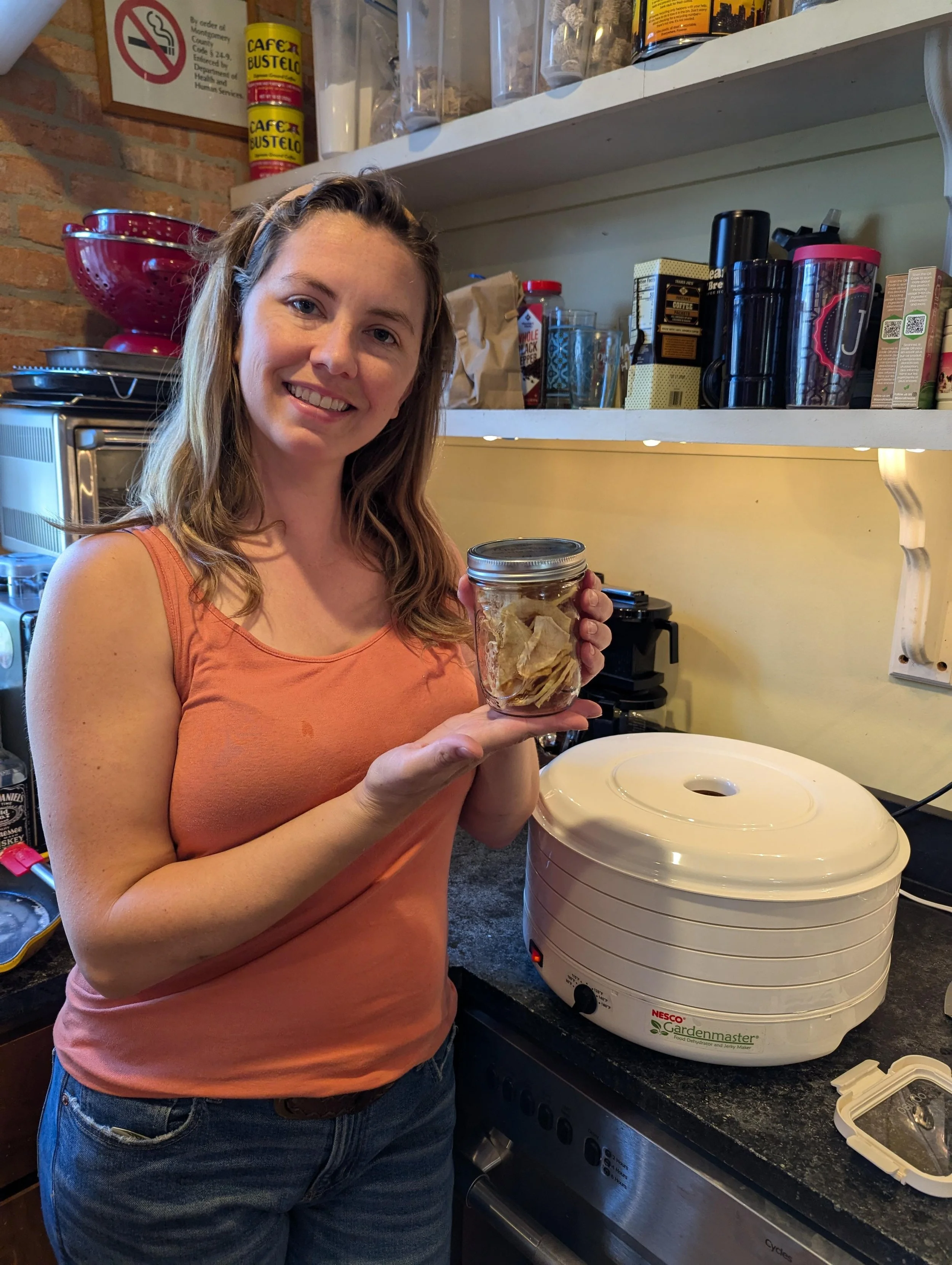 A woman standing in a kitchen, holding a glass jar filled with potato chips, with a food dehydrator and various kitchen items in the background.