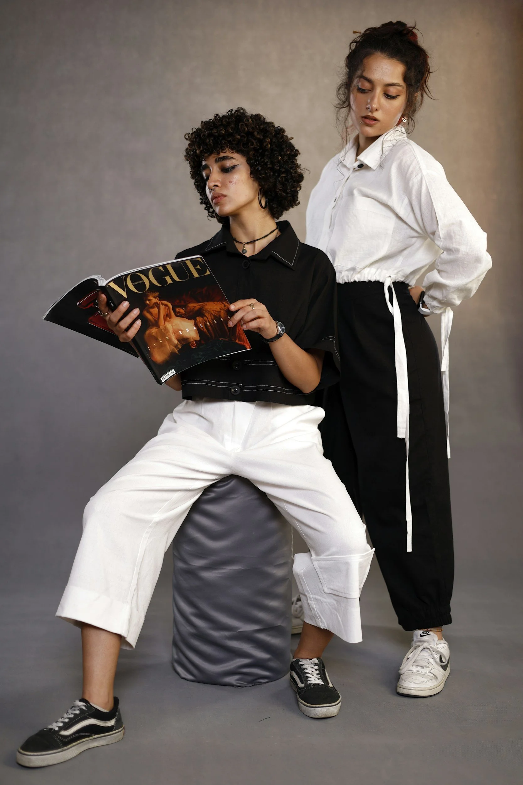 Two young women are posing against a neutral gray background. One woman is sitting on a stool with her legs apart, holding a Vogue magazine, wearing a black top and white wide-leg pants. The other woman is standing behind her, slightly to the side, wearing a white shirt and black pants with a white stripe, and has her hand on her hip.