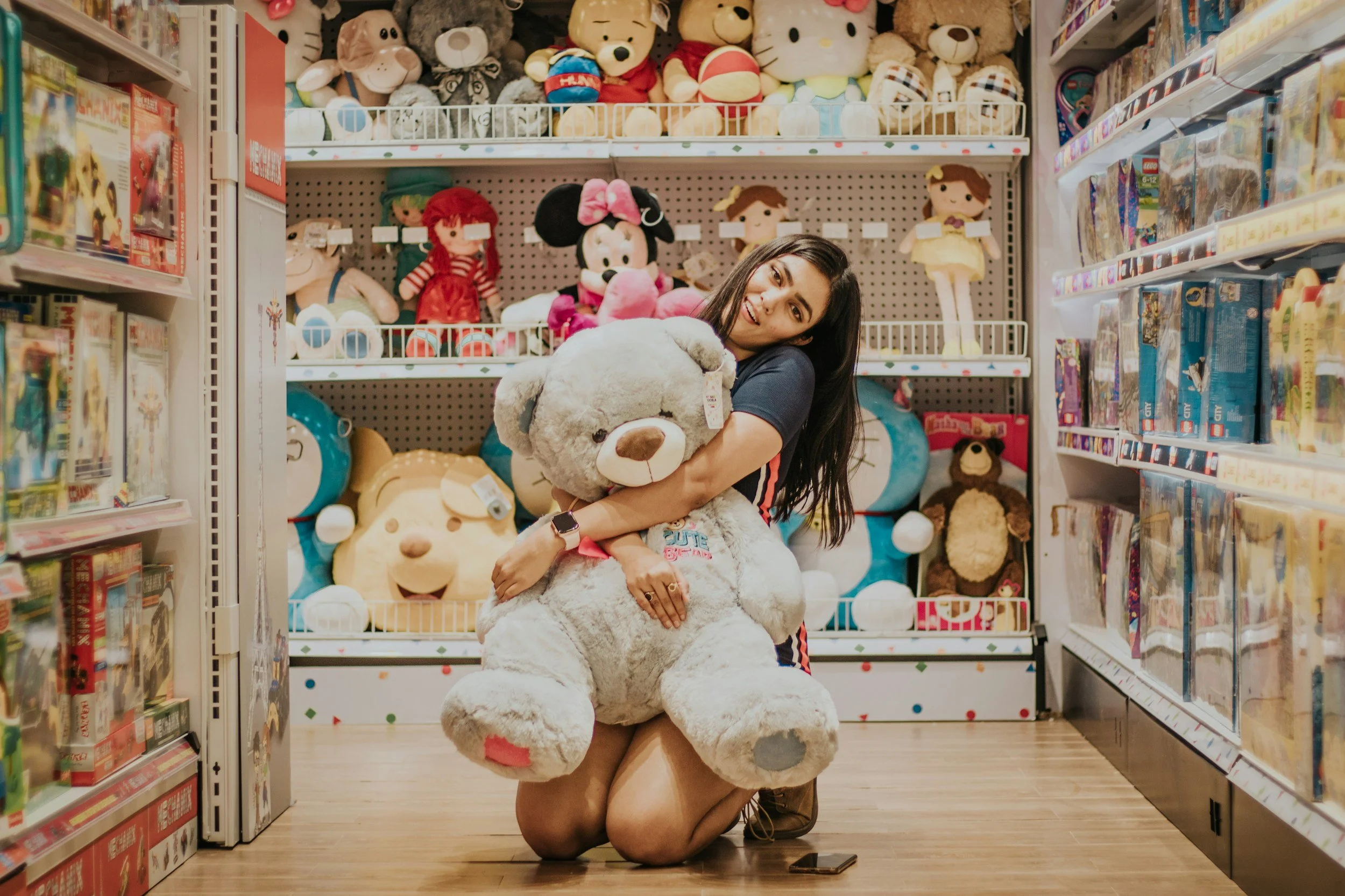 A woman kneeling on the floor of a toy store aisle, hugging a large plush teddy bear. Behind her, shelves are filled with various stuffed animals and toys, with a playful and colorful atmosphere.