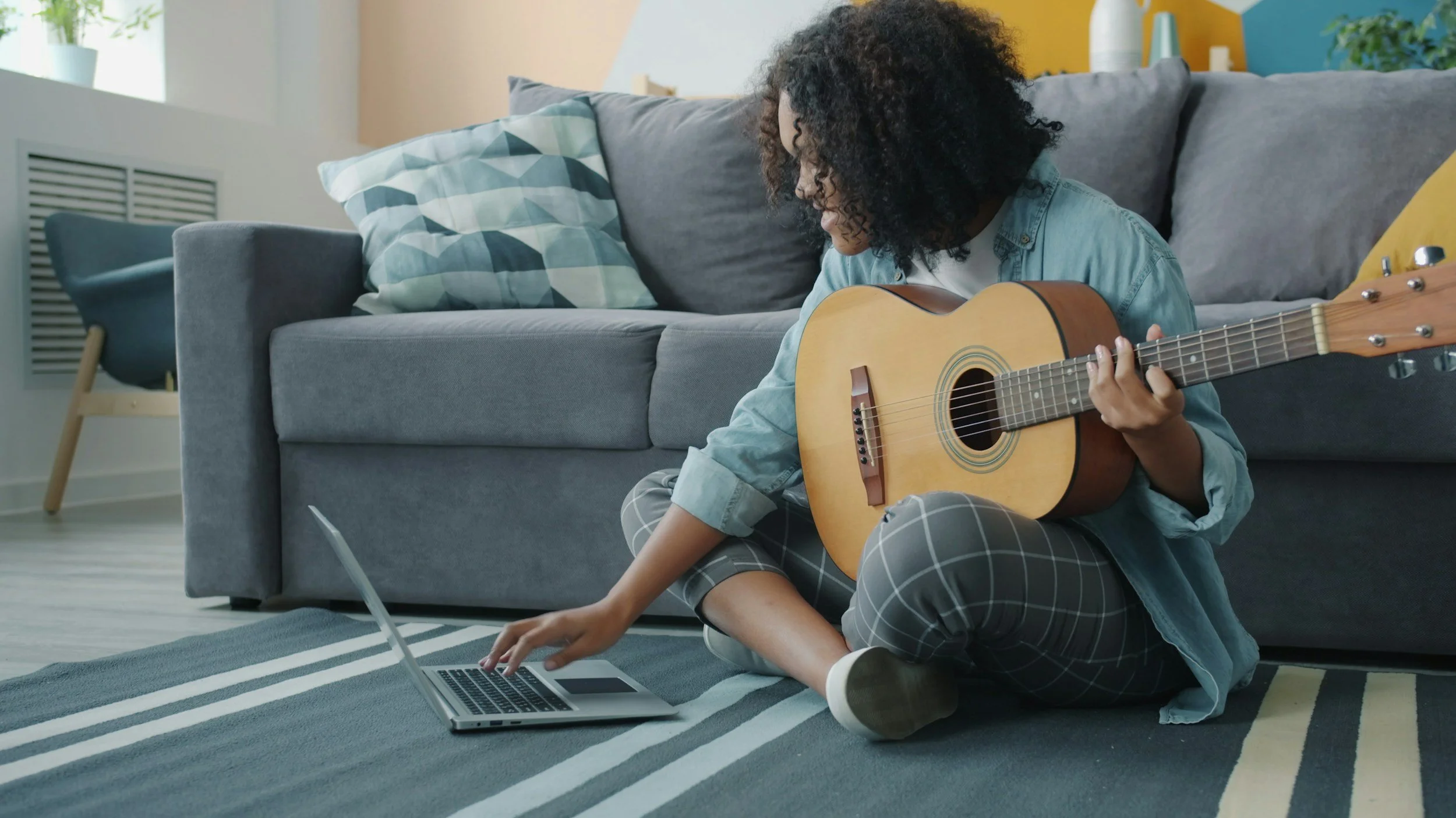 A person sitting on the floor in front of a laptop, using a guitar, in a living room with a gray couch, cushions, and a chair in the background.