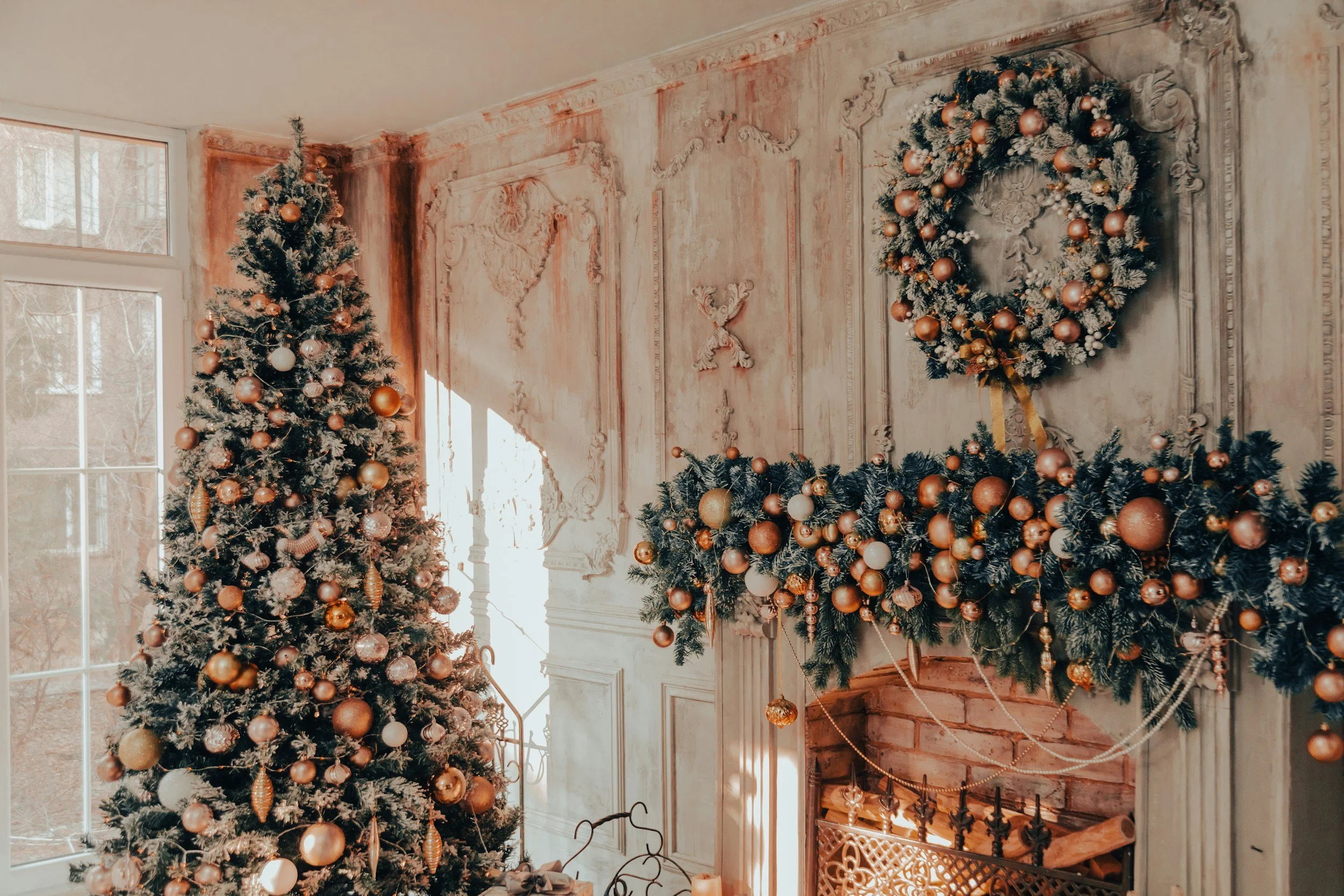 Decorated Christmas trees with ornaments and a gift box near a window in a room with ornate walls and a fireplace.