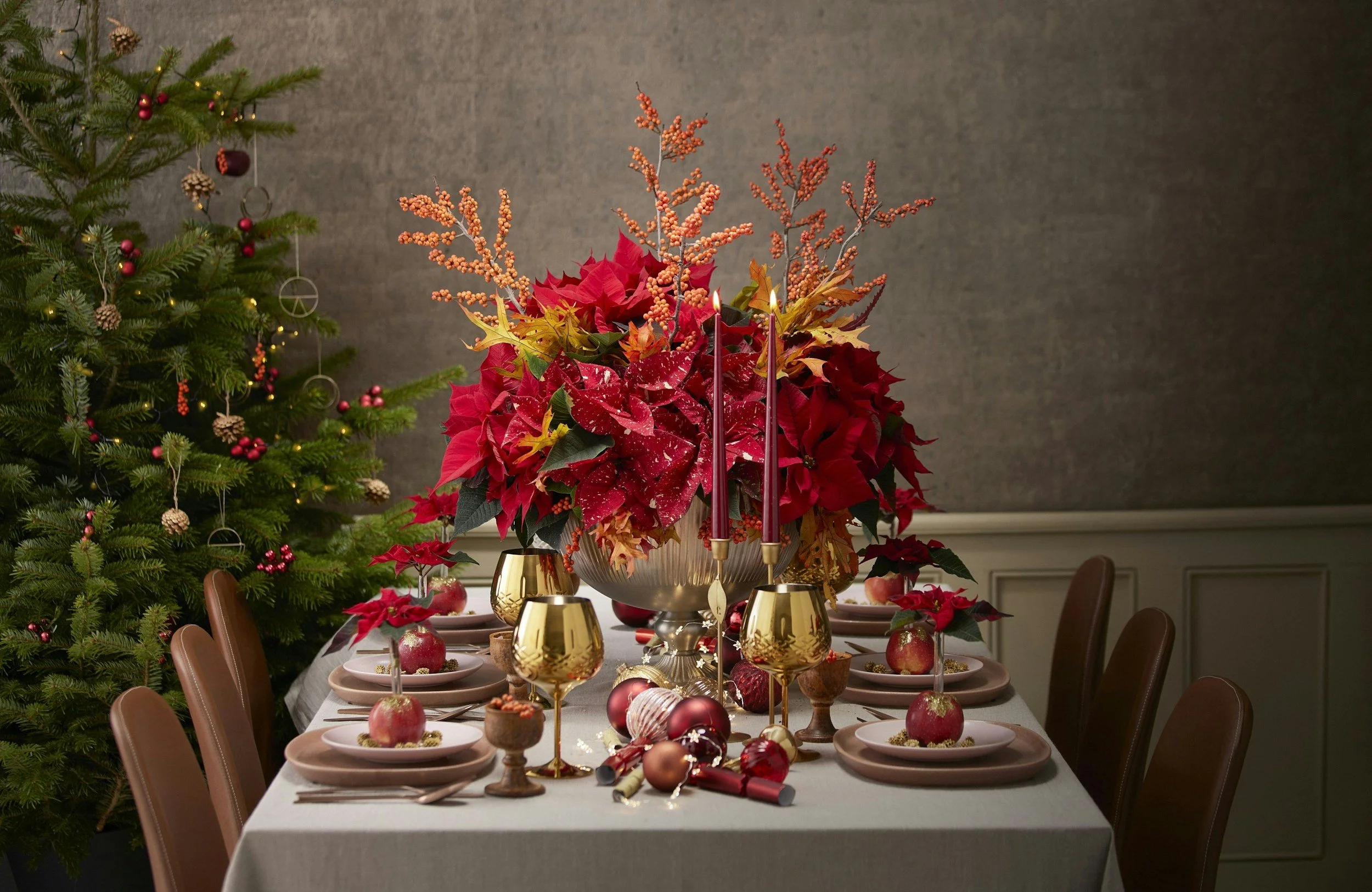 A decorated Christmas table with red and gold accents, surrounded by chairs, with a poinsettia centerpiece and a Christmas tree on the left decorated with ornaments and lights.