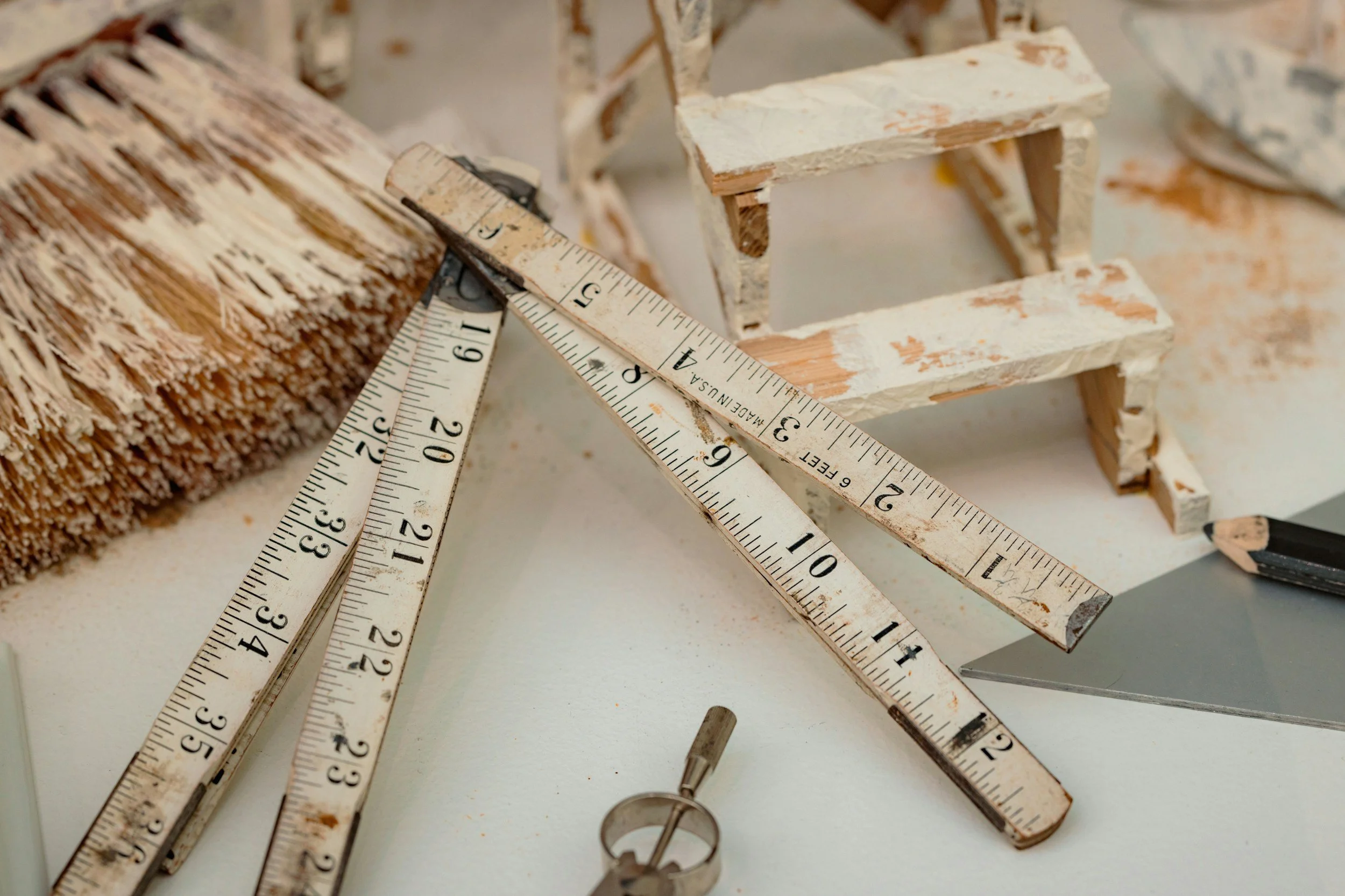 Close-up of a carpenter's workspace with a carpenter's square, a tape measure, and a wooden structure being painted with white primer.