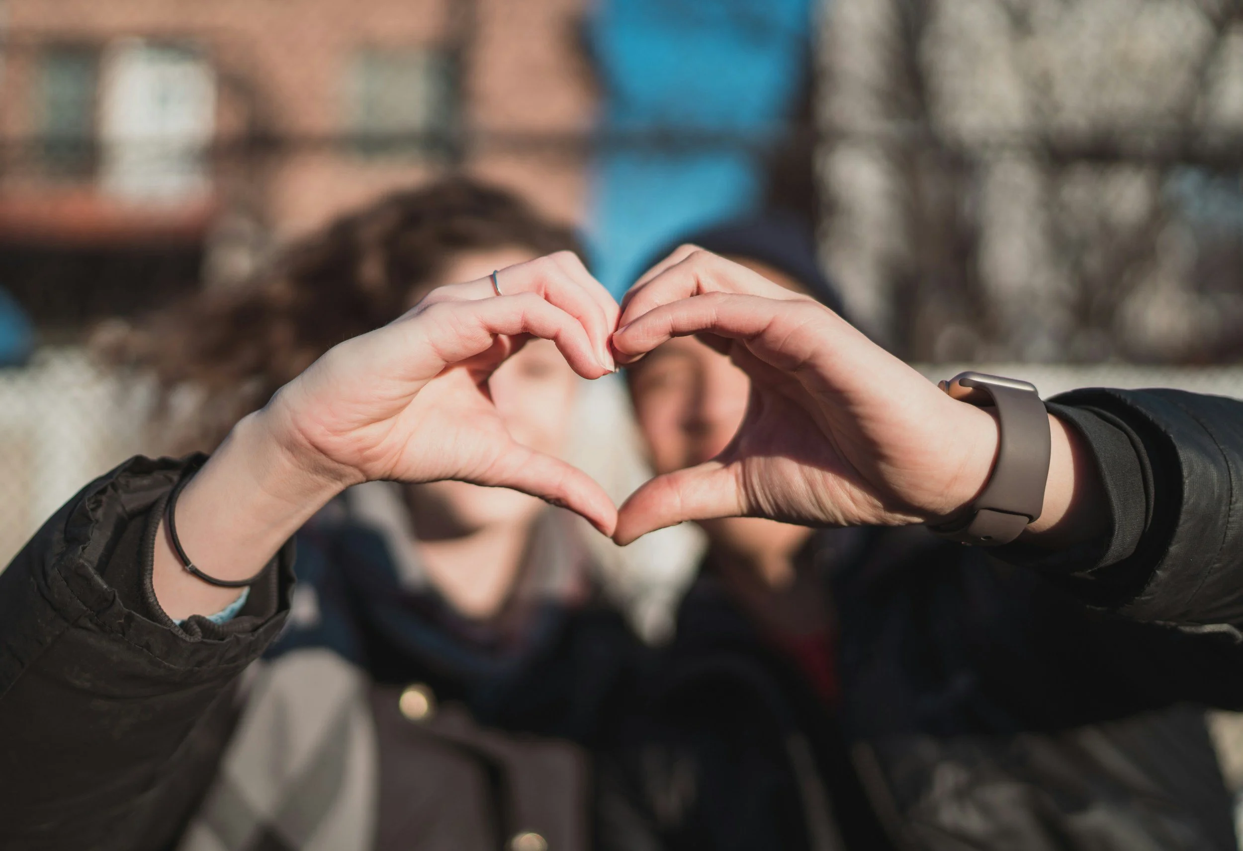 A person forming a heart shape with their hands outdoors, with a blurred background of other people and buildings.