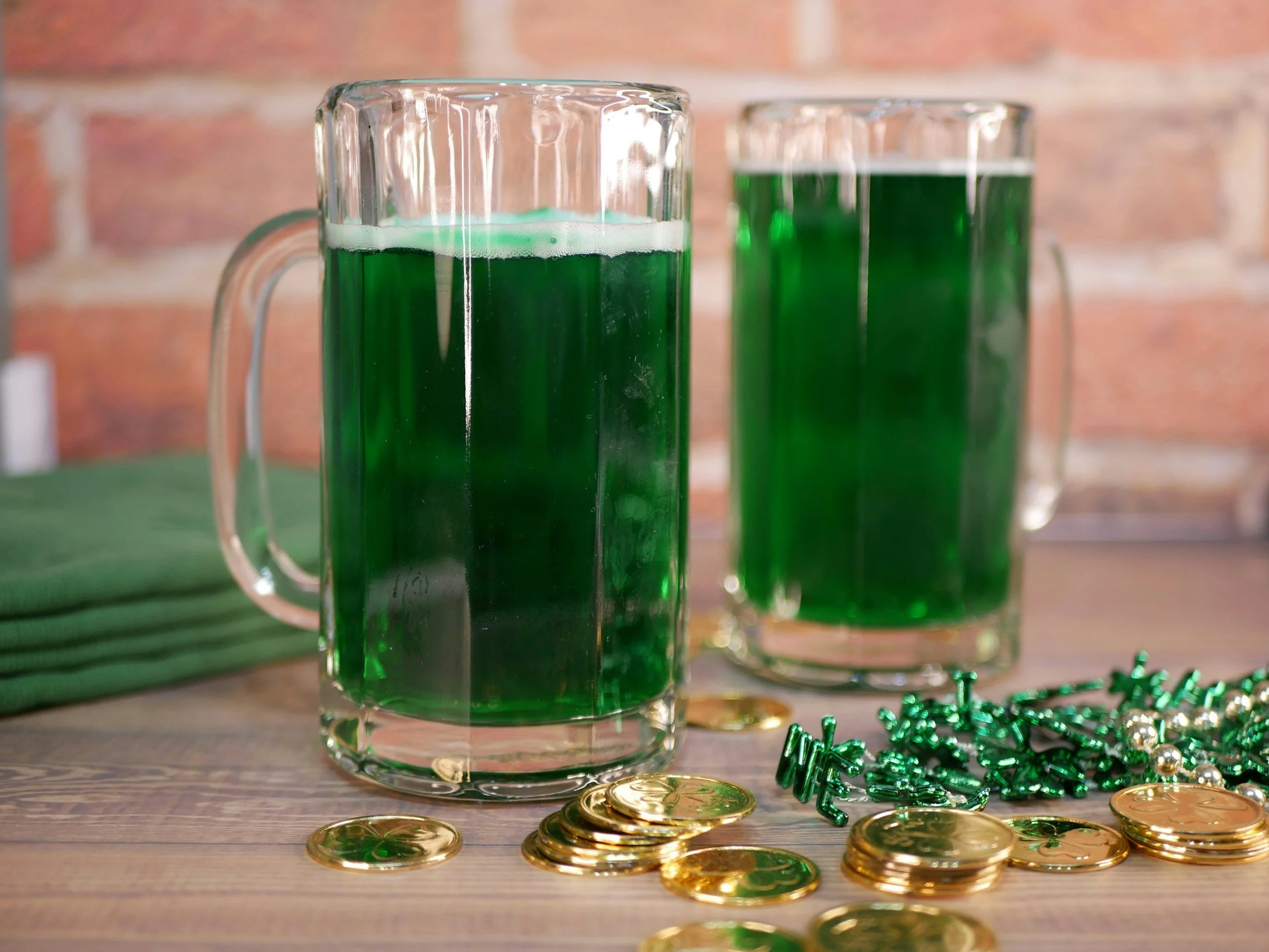A clear glass mug filled with green beverage, surrounded by scattered gold coins and green and gold decorative beads on a wooden surface, with a blurred brick wall background.