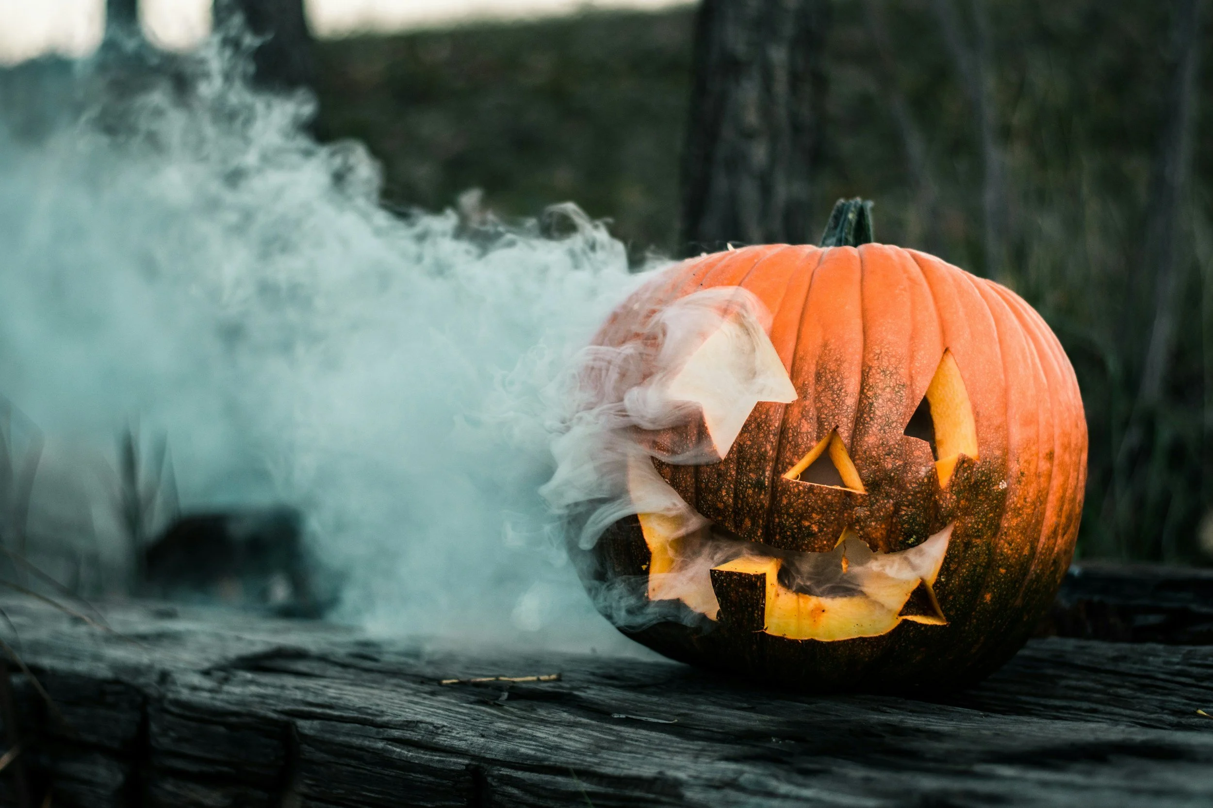 A carved pumpkin with a smiling face sits on a dark wooden surface, emitting smoke or fog from its top, with a blurred outdoor background.