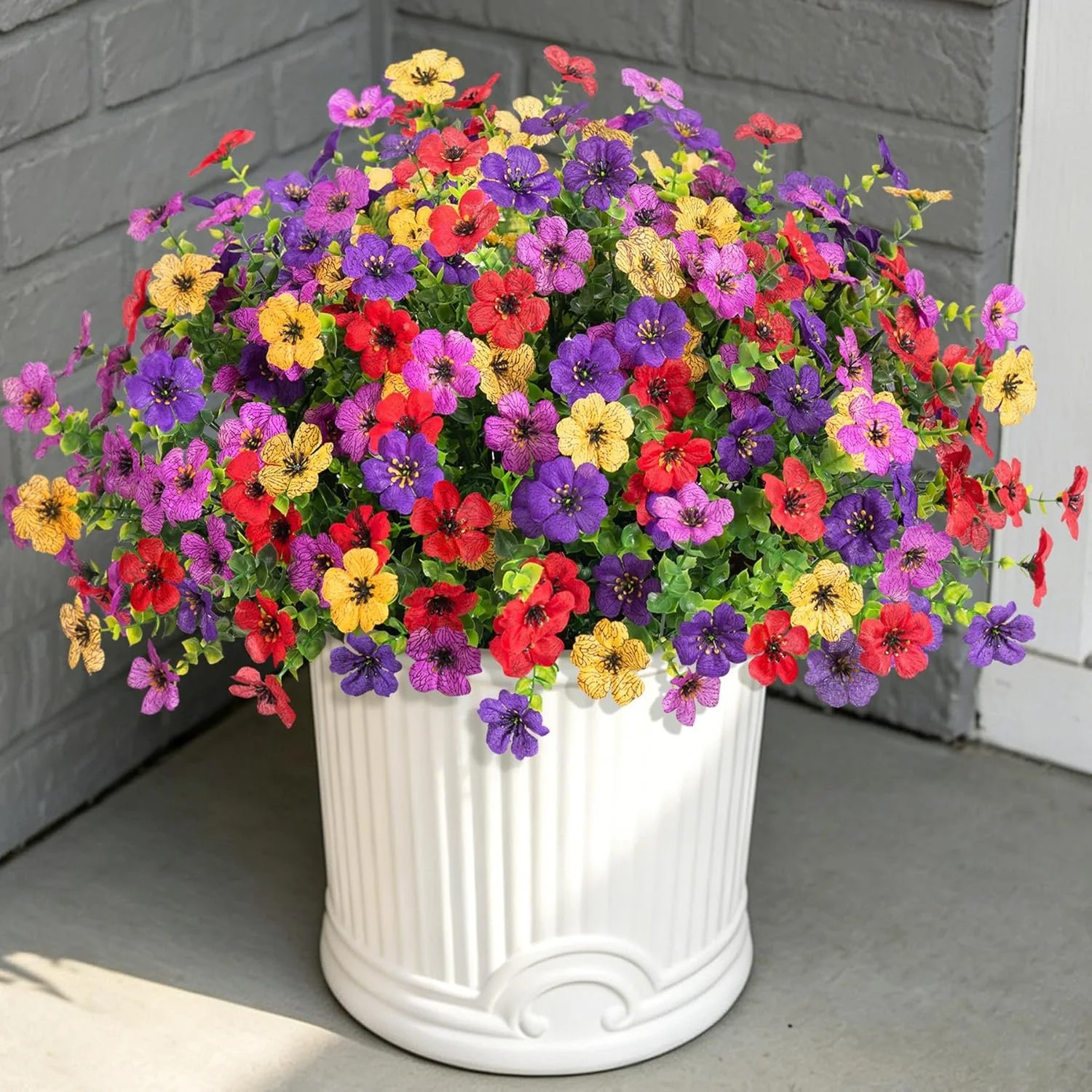 Colorful pot of small purple, yellow, and red flowers with green leaves on a porch, against gray brick wall