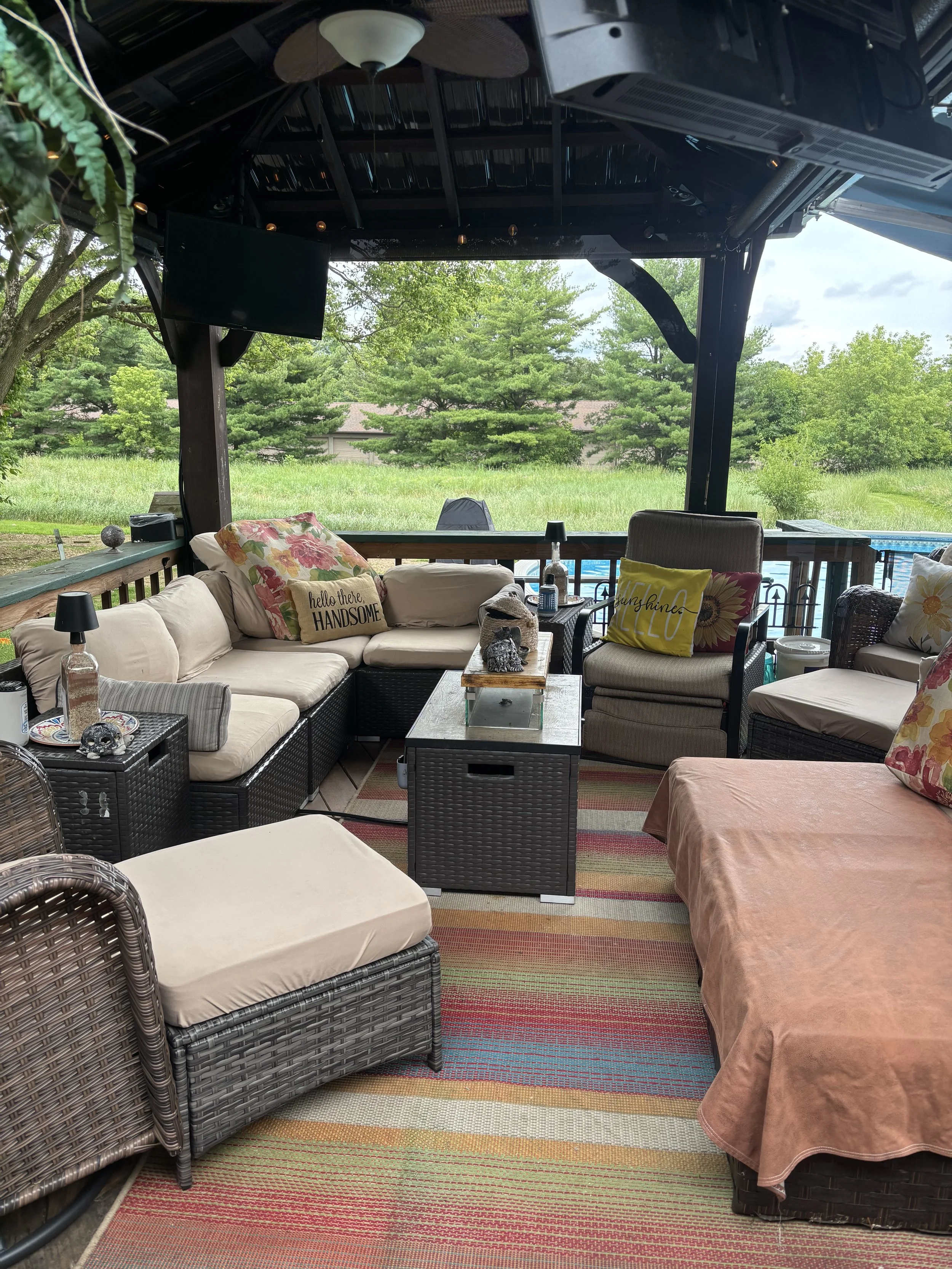 Outdoor patio with wicker and fabric furniture, cushions, a TV, and a view of trees and grass.