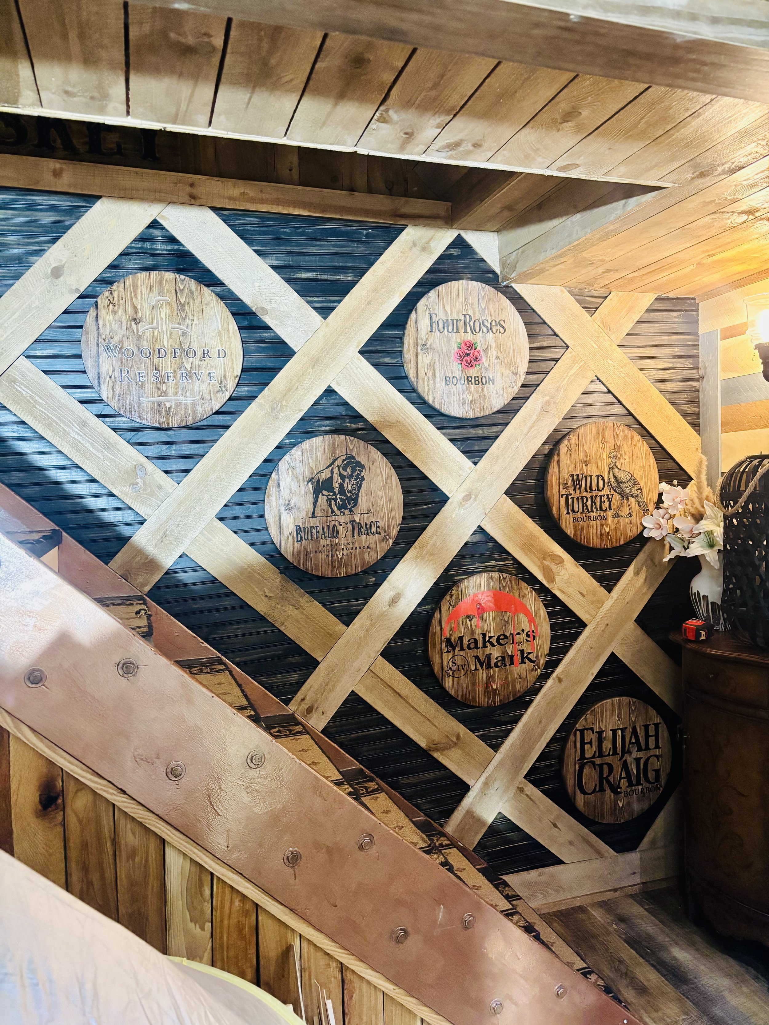 Decorative wooden wall with circular signs displaying bourbon brand names, including Woodford Reserve, Four Roses, Buffalo Trace, Wild Turkey, Maker's Mark, and Elijah Craig, mounted on a crisscrossed wooden frame.